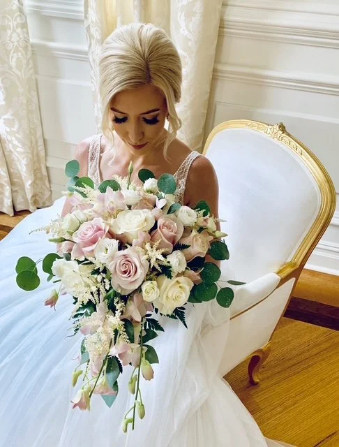 A bride with blonde hair holding a bouquet of pink and white roses and greenery, sitting on a white and gold elegant chair in a room with cream-colored curtains.