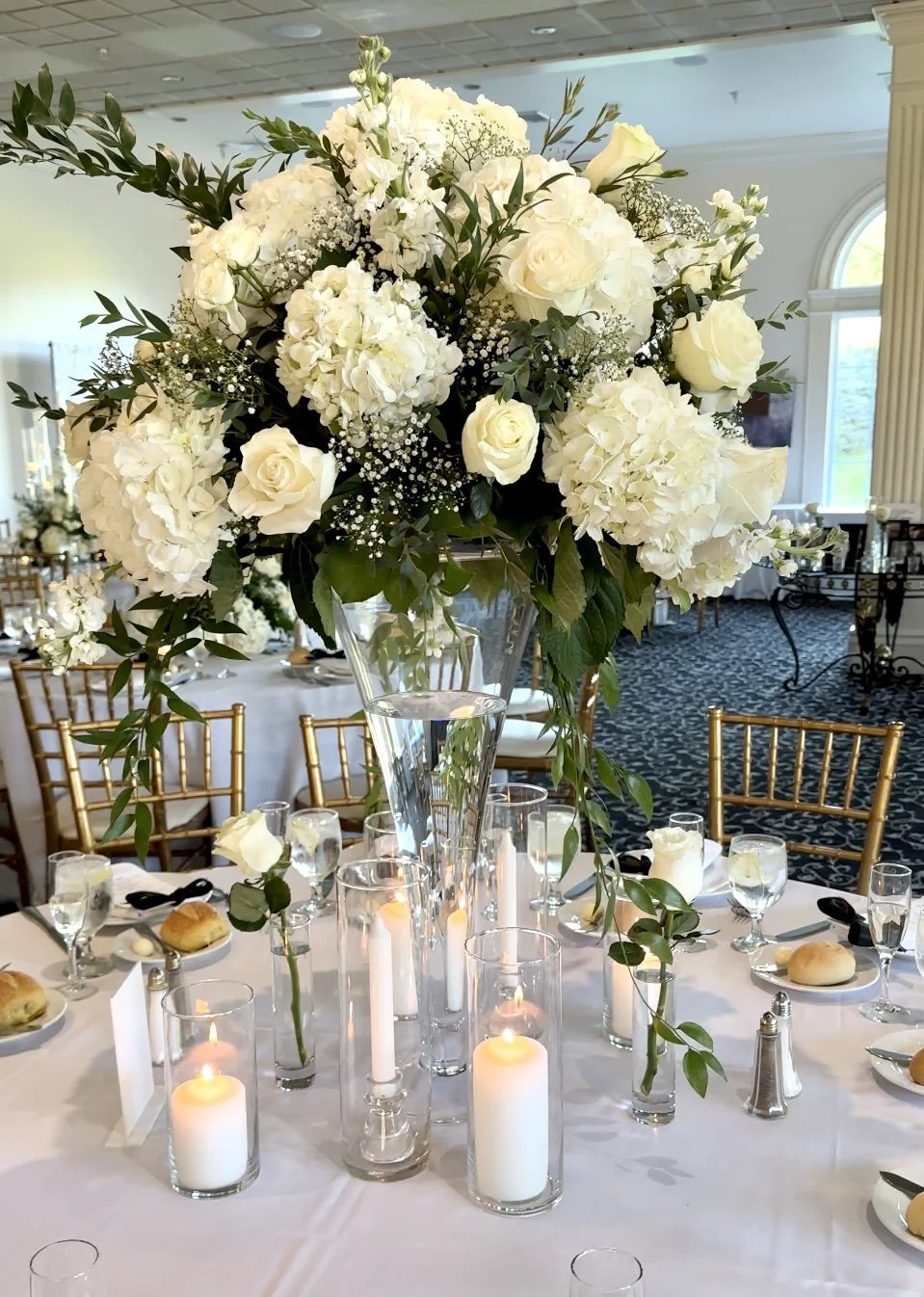 A large white flower arrangement with roses, hydrangeas, baby's breath, and greenery in a tall glass vase, placed on a dinner table decorated with candles and plates in a decorated banquet hall.