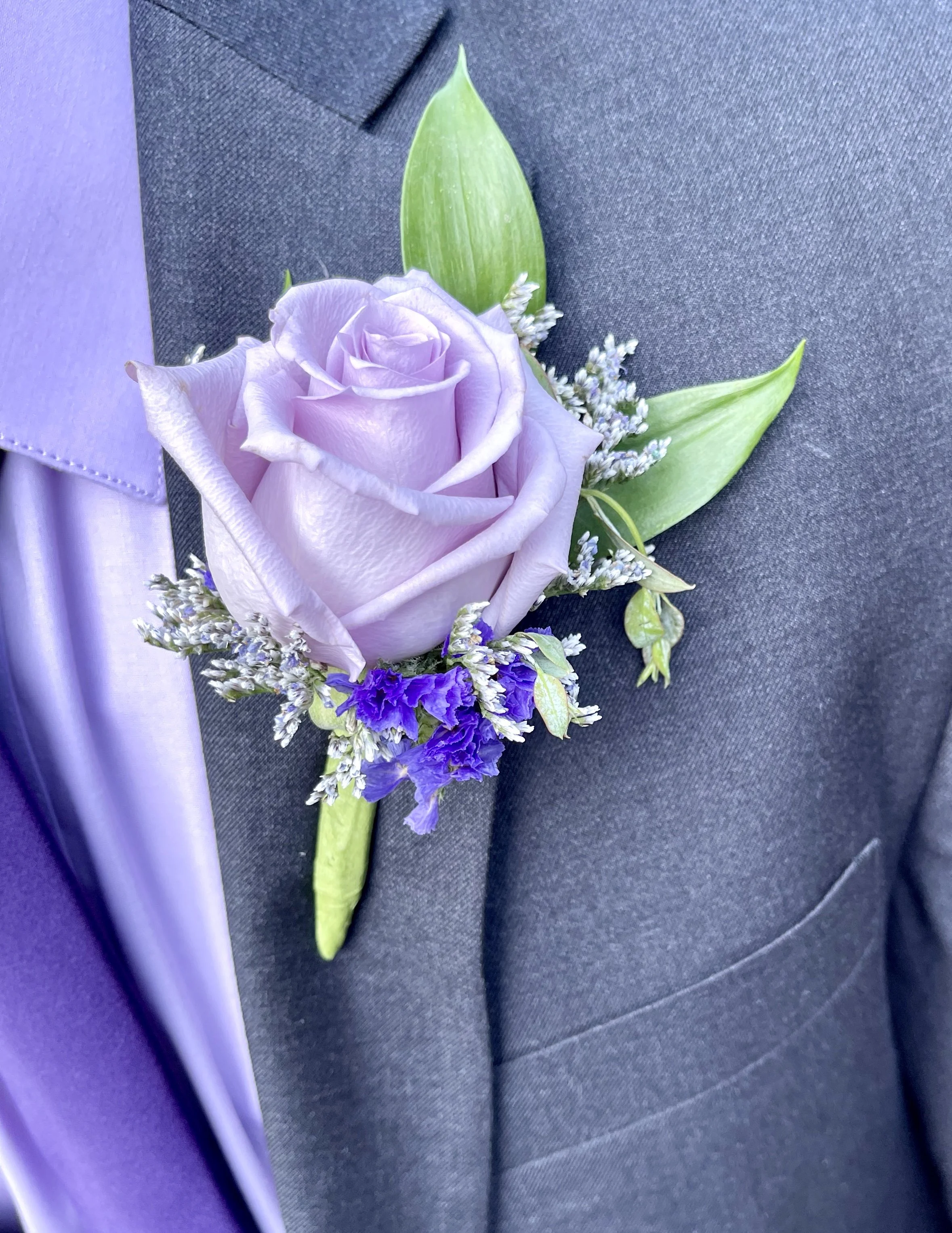 A boutonniere with a lavender rose, white and purple small flowers, and green leaves pinned to a dark suit fabric.
