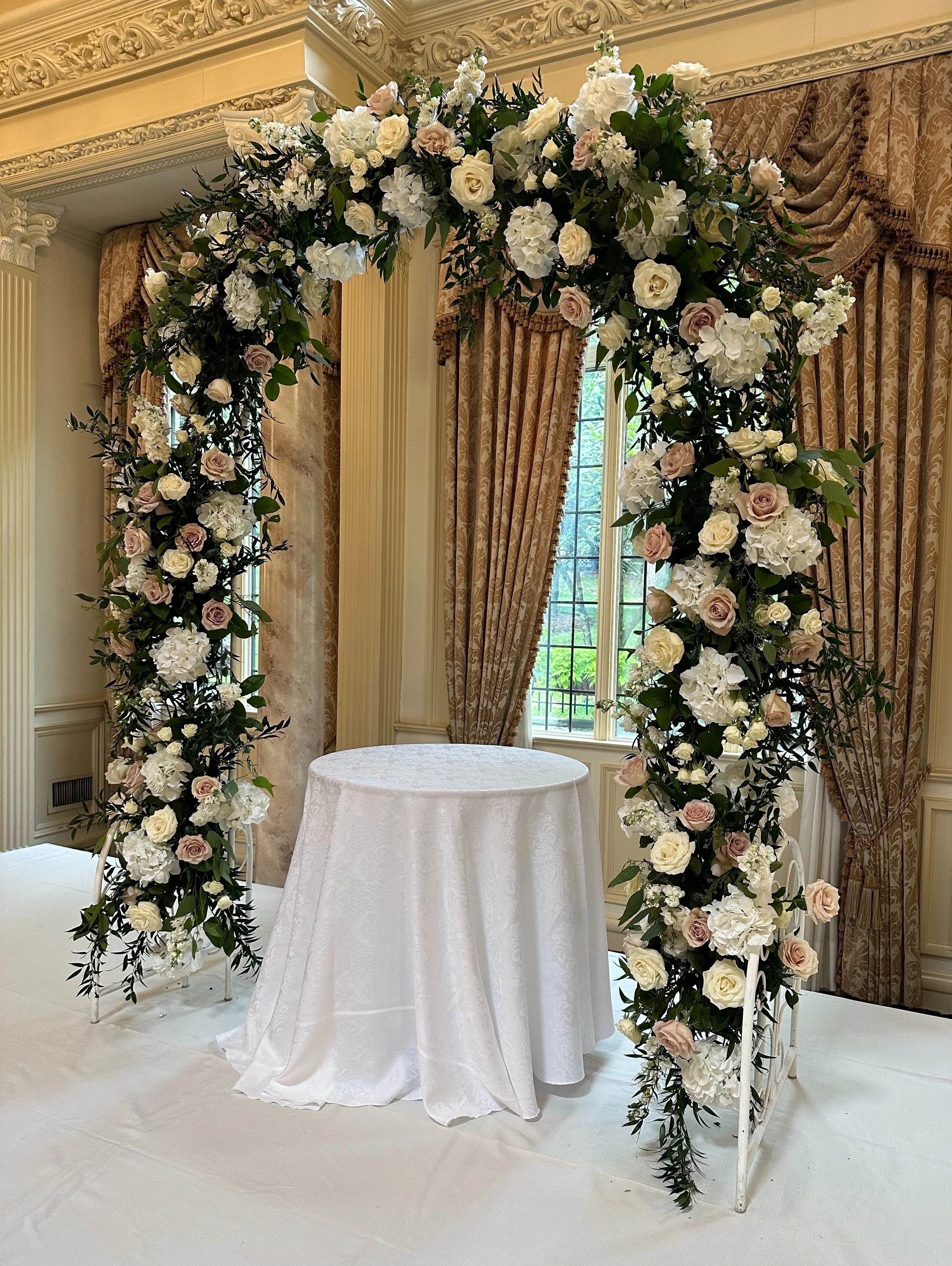 A decorated wedding arch made of white metal, adorned with white and pale pink roses, hydrangeas, and greenery, set in an elegant room with beige patterned curtains and large windows.