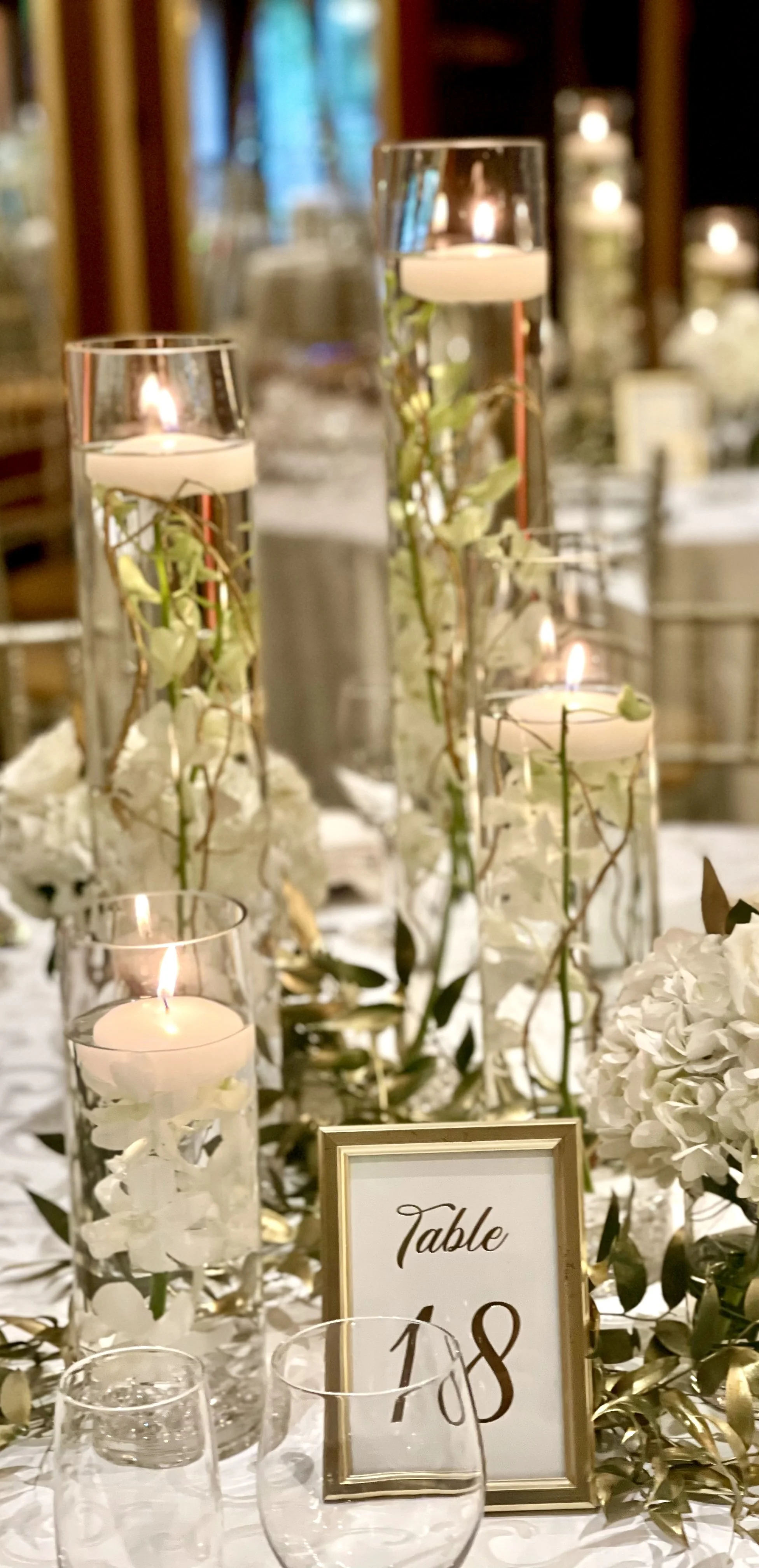 Elegant wedding reception table centerpiece with white flowers, candles in tall glass holders, and a table number sign reading 'Table 18'.