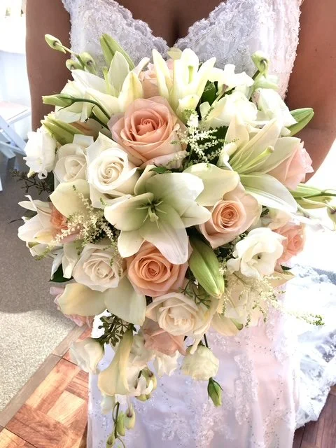 A bride holding a cascading bouquet of white and light pink roses, white lilies, and greenery, in a wedding dress.