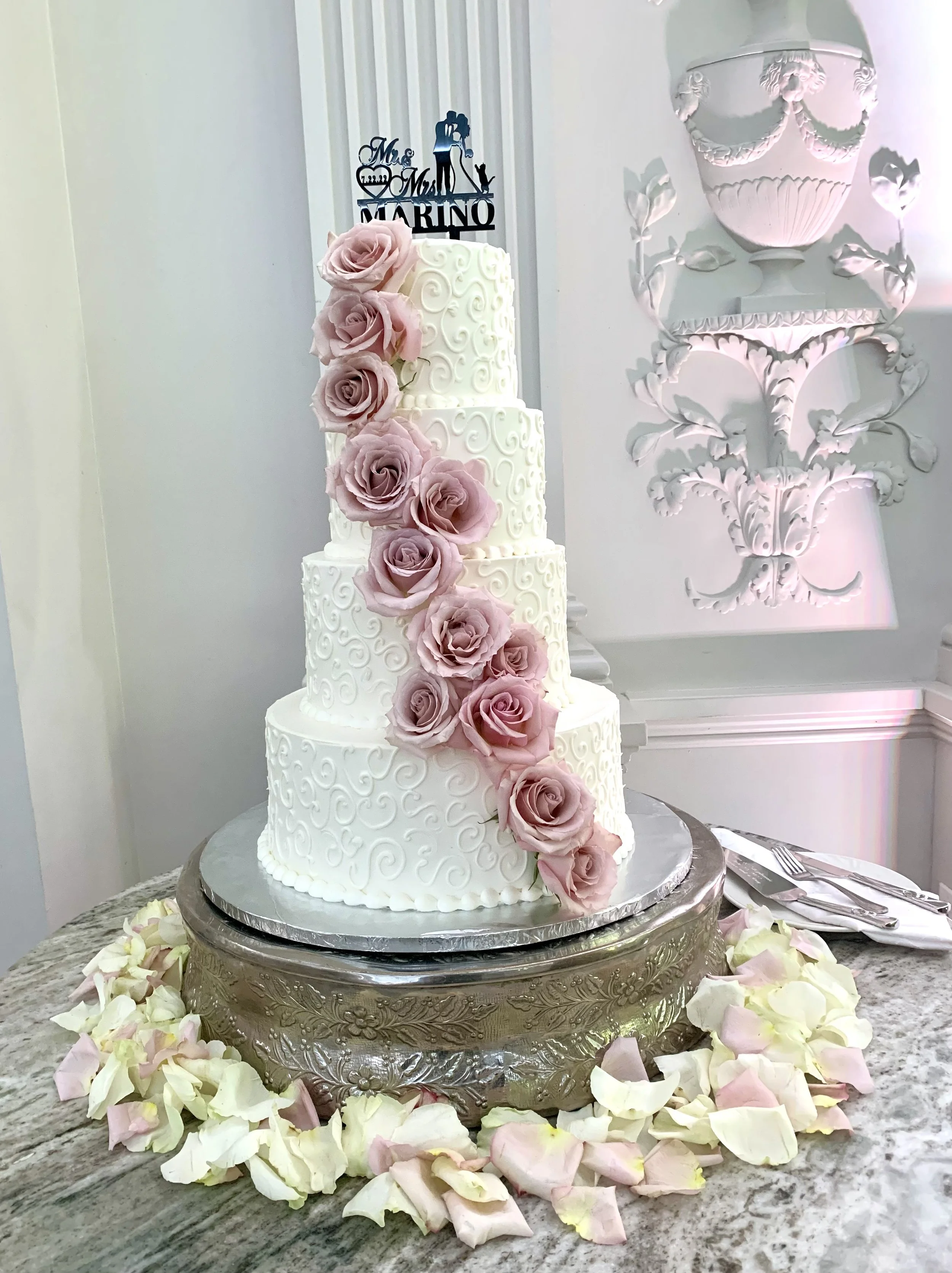 A multi-tiered wedding cake decorated with cascading pink roses and intricate white frosting swirl patterns, placed on an ornate silver cake stand surrounded by scattered white and pink rose petals, with a decorative wall sculpture of a urn and flowe