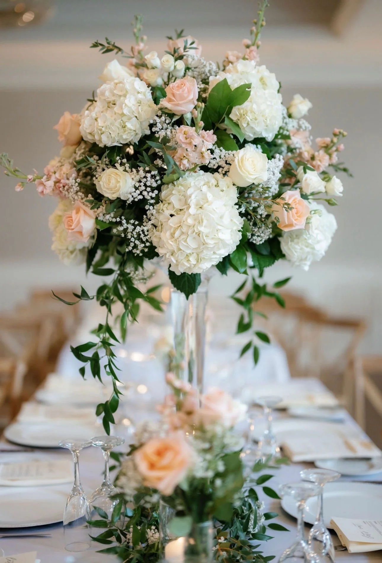 A floral centerpiece with white hydrangeas, pale pink roses, and green foliage on a table, set for a formal event or wedding reception.