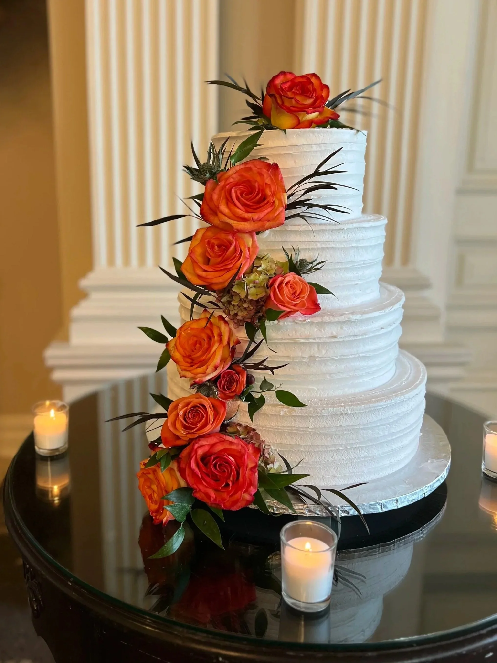 A multi-tiered white wedding cake decorated with orange and pink roses and green foliage, placed on a black table with candles around it.