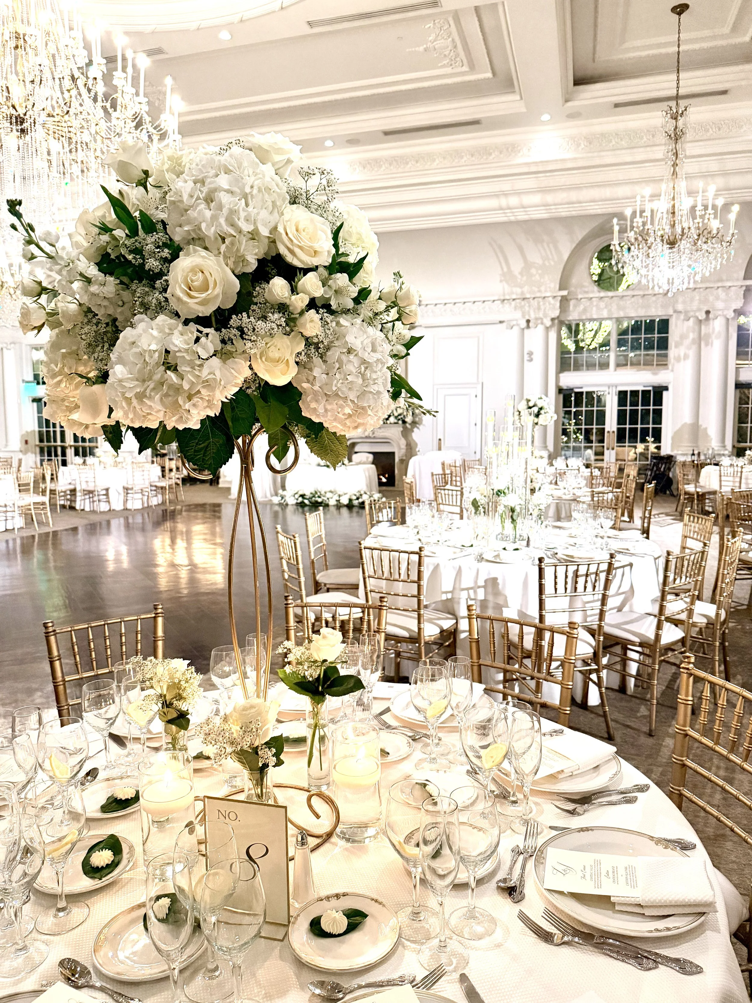 Elegant wedding reception hall decorated with white flowers, gold chairs, chandelier lighting, and table settings.
