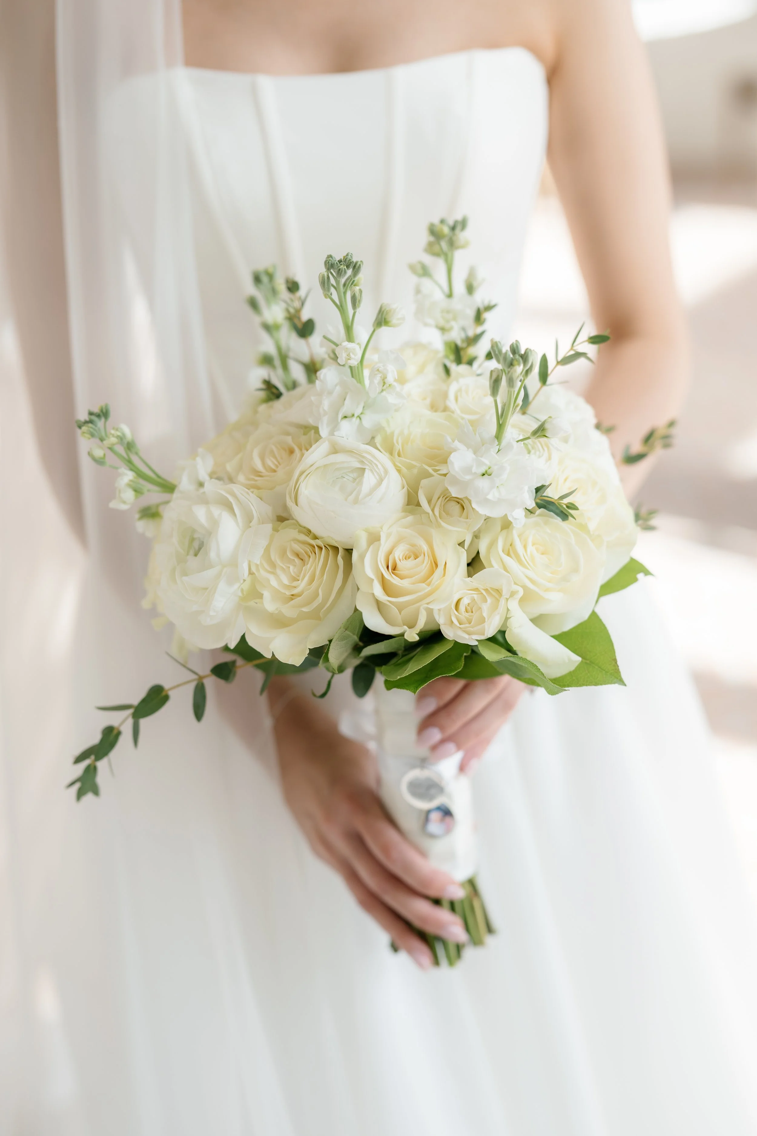 A bride holding a bouquet of white roses and greenery, wearing a strapless white wedding dress.