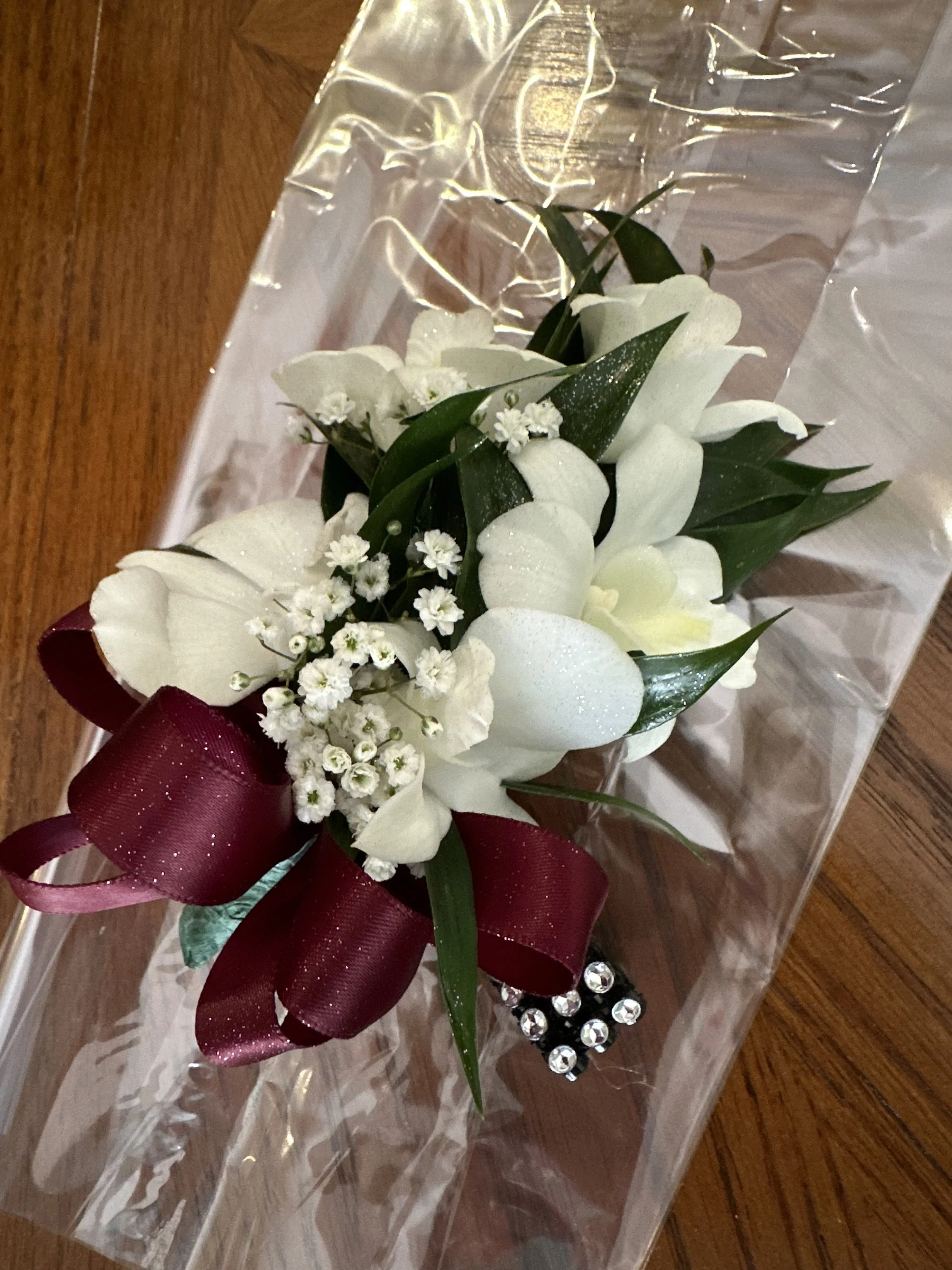 A bouquet of white flowers, small white blossoms, and green leaves wrapped with a red ribbon, inside a clear plastic wrapper with decorative rhinestones, on a wooden surface.