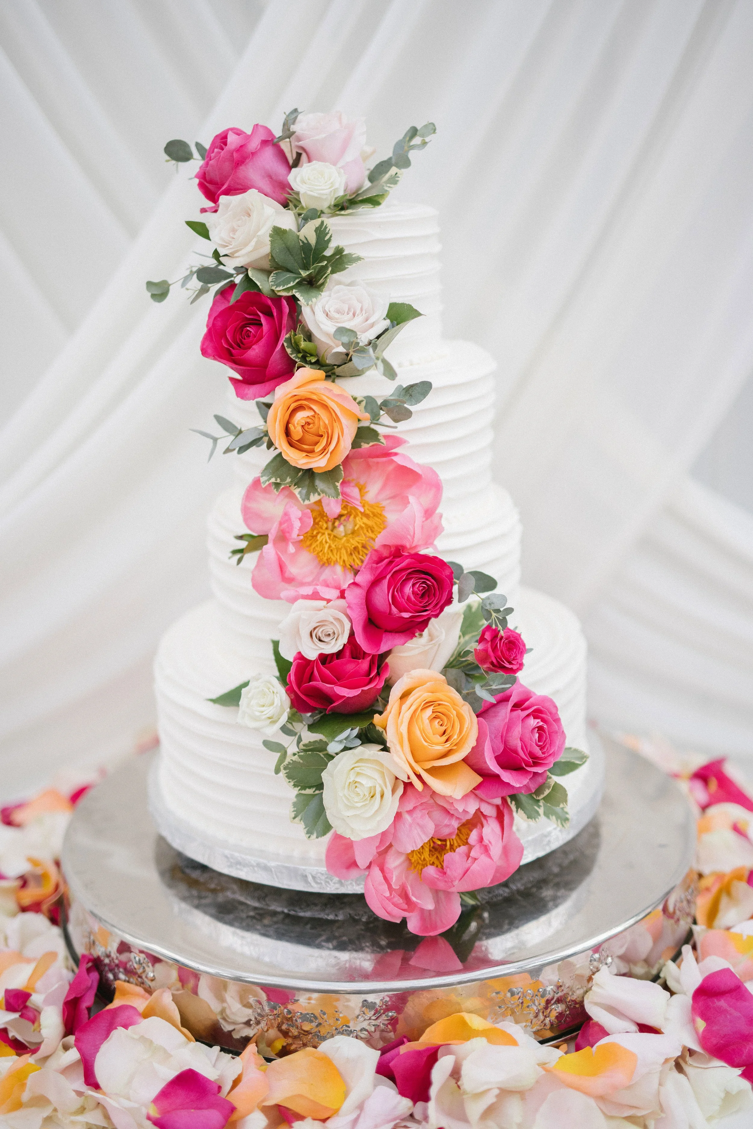 A multi-tiered white wedding cake decorated with various pink, peach, and white roses, along with greenery, sitting on a stand surrounded by scattered flower petals.