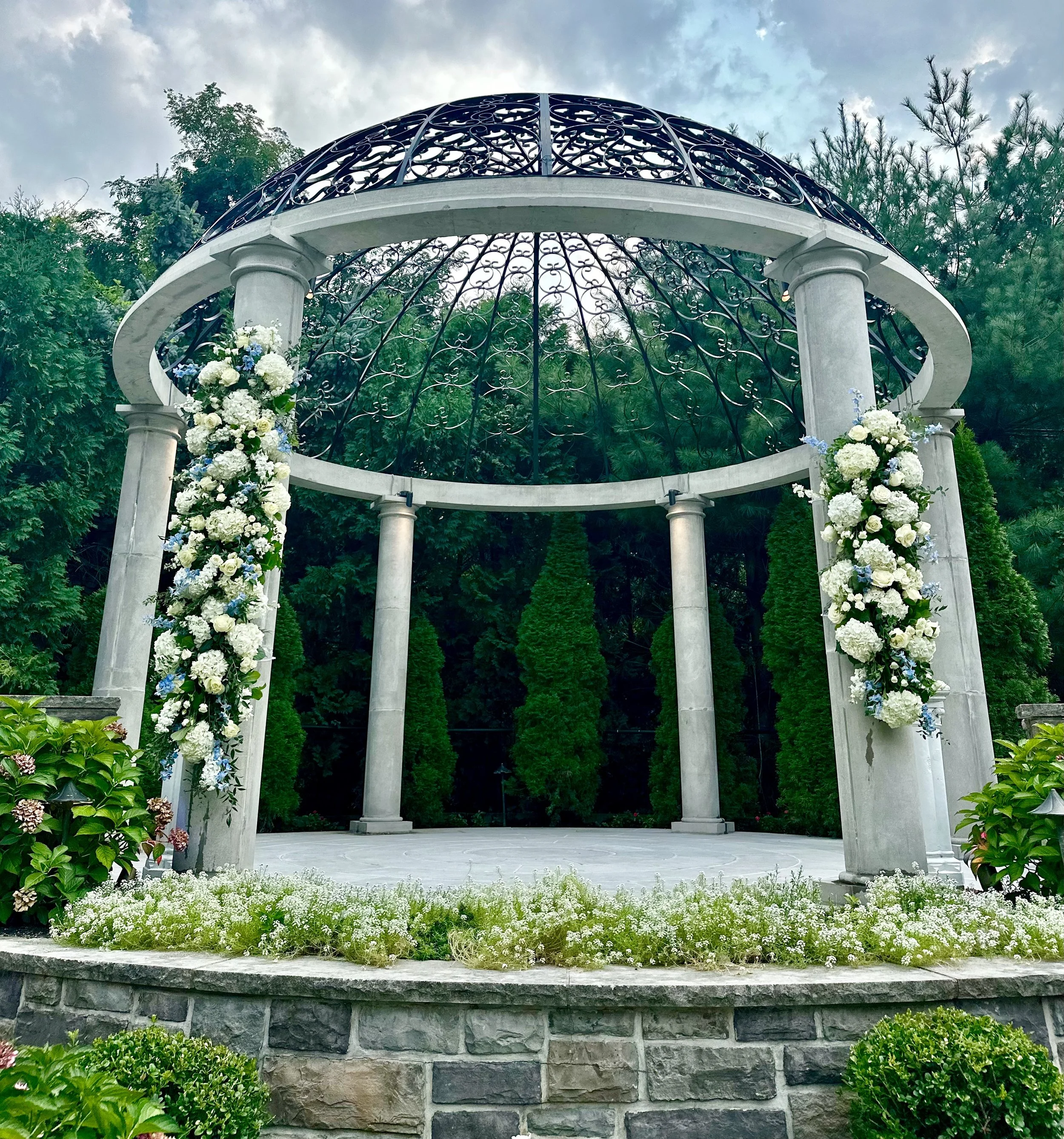 A white round gazebo with columns, decorated with white and blue flowers, situated in a garden with bushes and trees, under a cloudy sky.