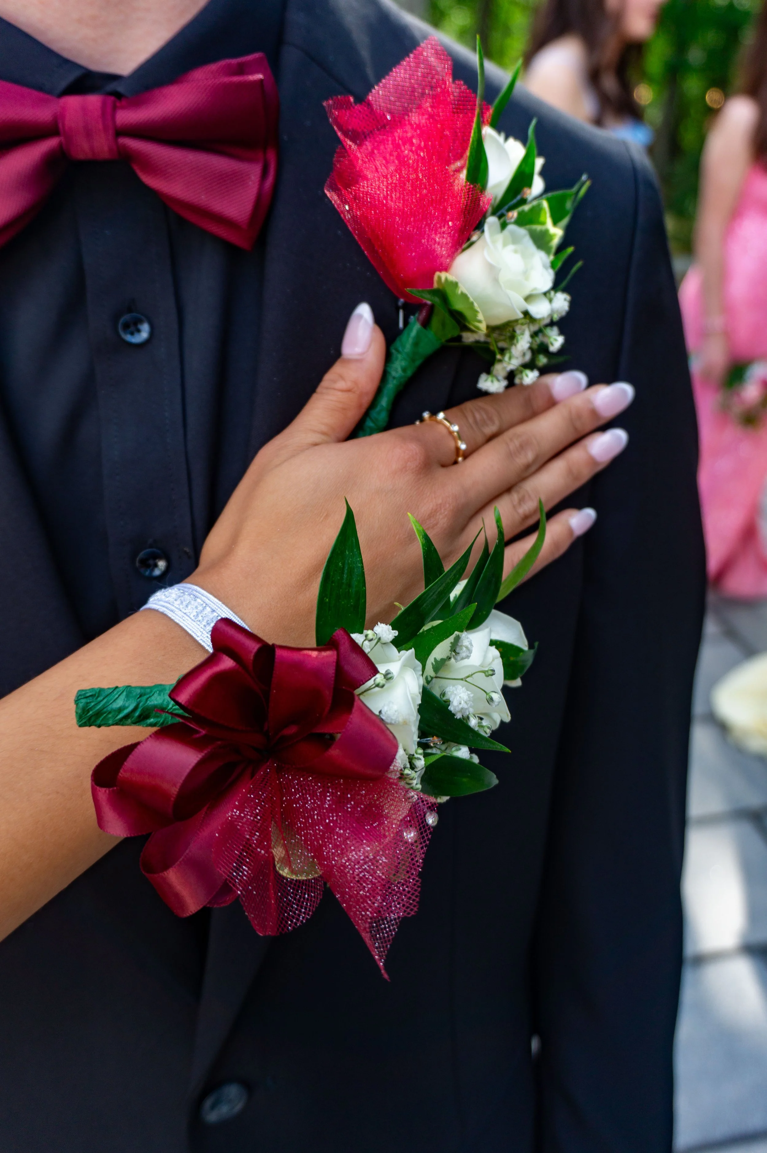A person wearing a black suit with a red bow tie and a floral boutonniere featuring red, white, and green flowers, with a woman in a pink dress in the blurred background.