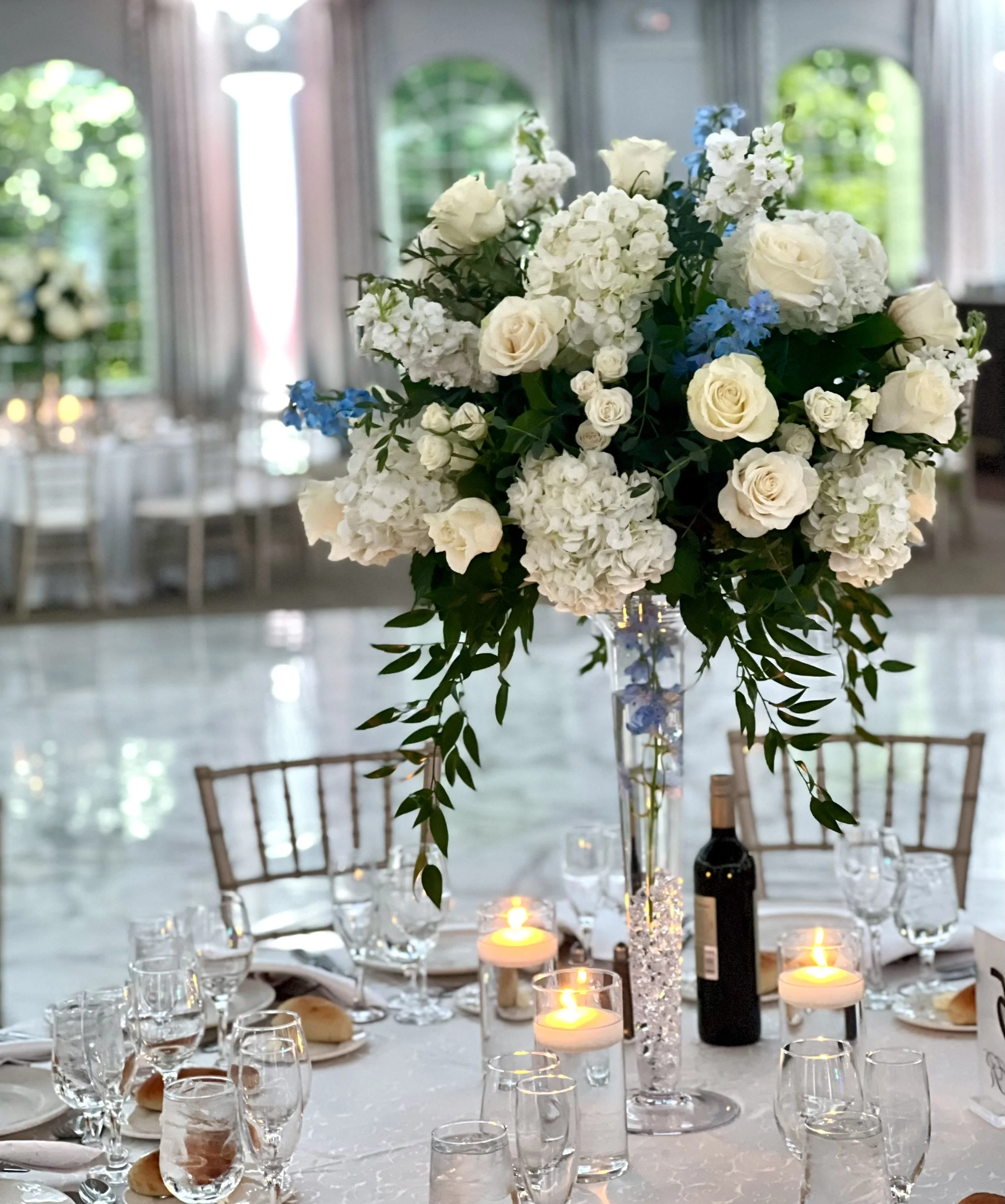 A table set for a formal event with a large floral centerpiece consisting of white roses, hydrangeas, and small blue flowers, surrounded by lit candles in small vases, glassware, and bread rolls, with chairs and windows in the background.