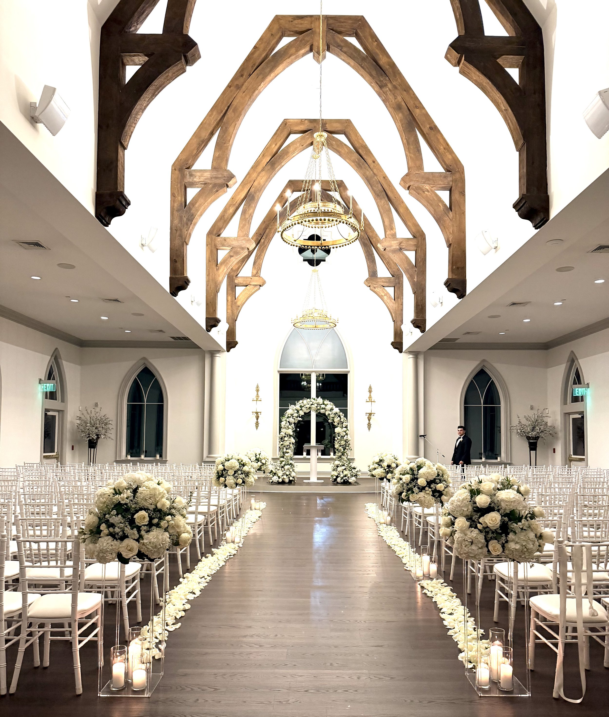 Elegant wedding ceremony setup inside a church with rows of white chairs decorated with white flowers, a floral arch at the altar, and candles lining the aisle.