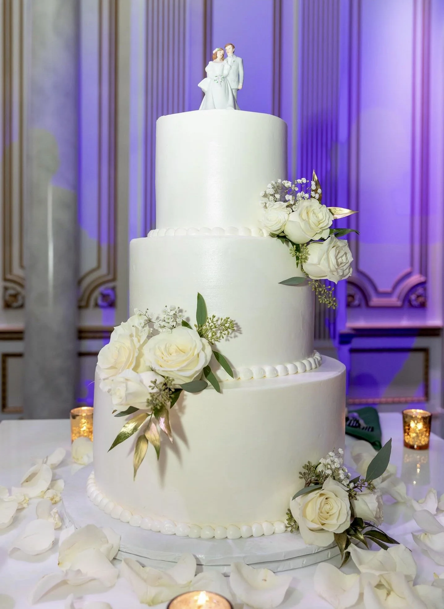 A white wedding cake decorated with white roses, greenery, and gold accents, with a bride and groom topper, surrounded by rose petals and candles.