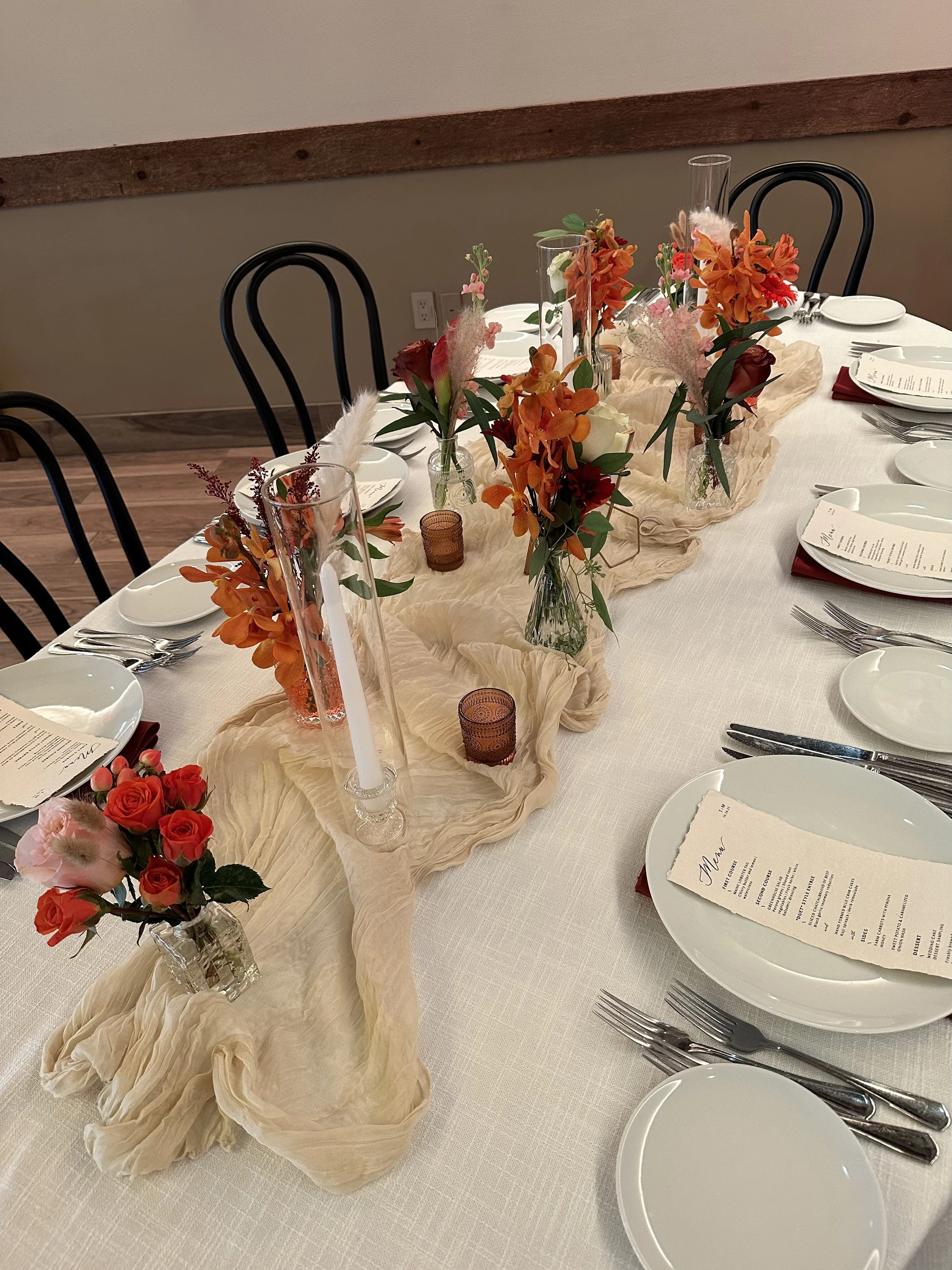 A decorated dining table with a white tablecloth, floral centerpieces in clear glass vases, tall candles, and place settings that include white plates, silverware, and menus. The decor features orange, pink, and red flowers along with soft fabric acc