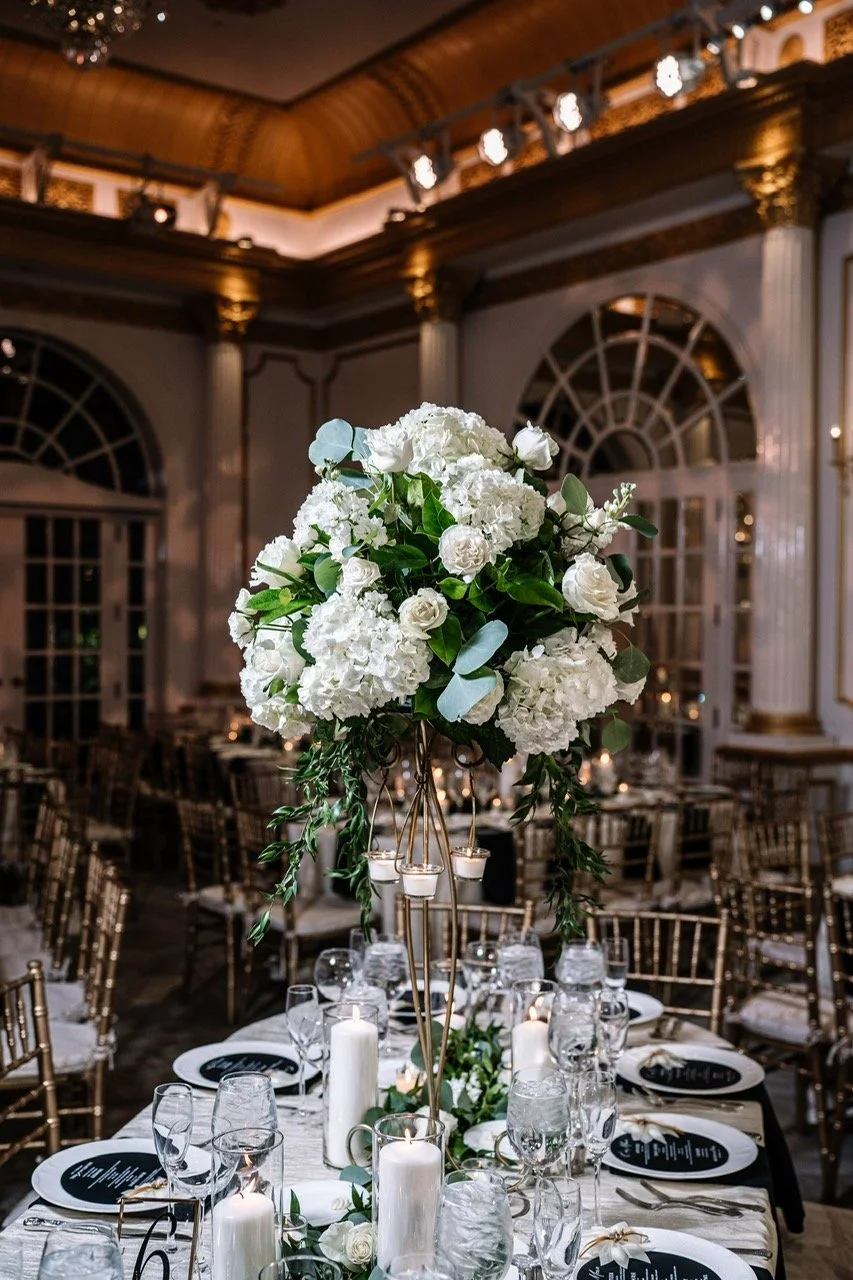 Elegant wedding reception table with tall floral centerpiece of white hydrangeas and roses, surrounded by candles and glassware in a grand, ornate hall.