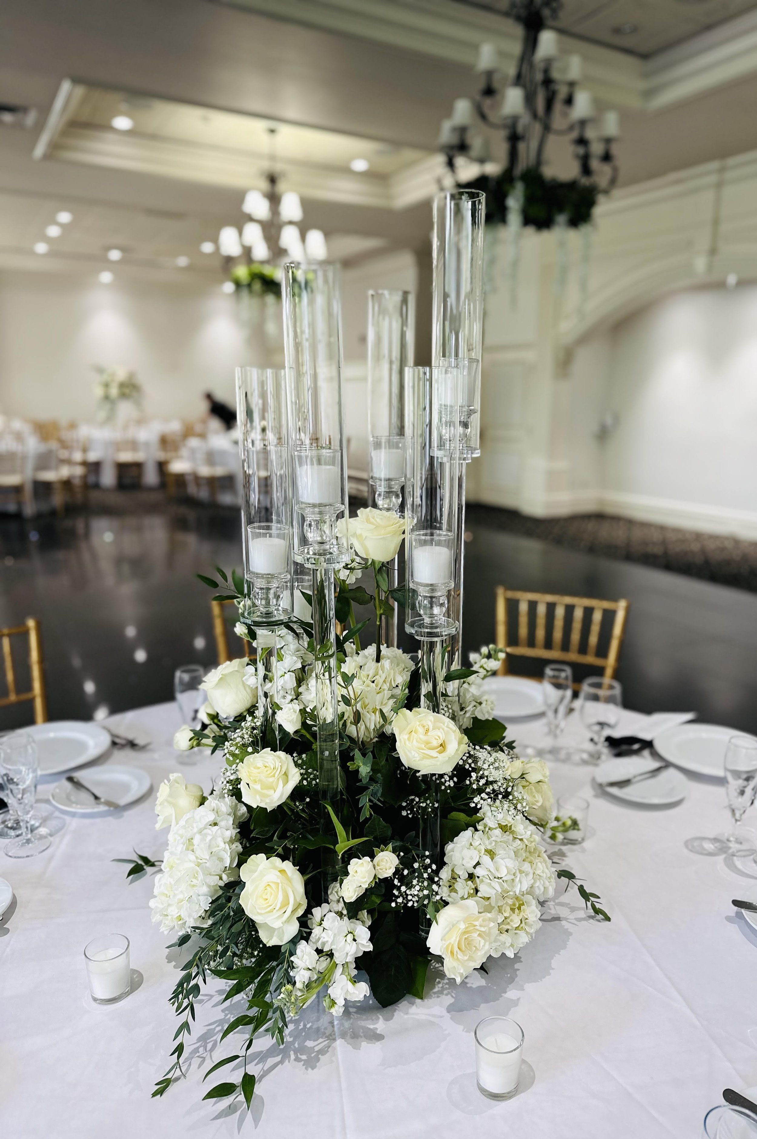 Elegant floral centerpiece with white roses, hydrangeas, and candles on a round wedding reception table with white tablecloth and gold chairs.