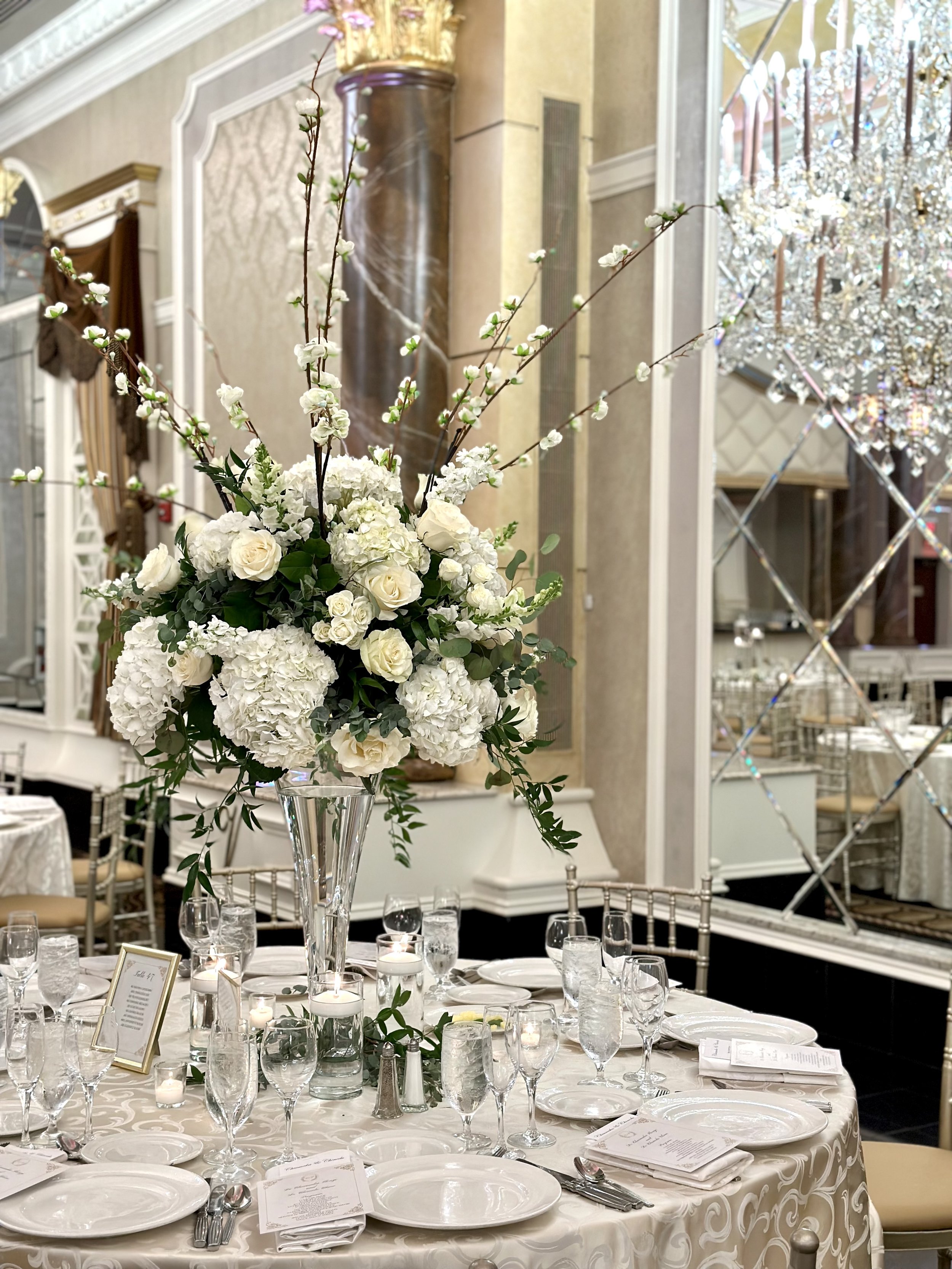 A large floral centerpiece with white roses, hydrangeas, and cherry blossom branches in a tall glass vase on a decorated banquet table at a wedding reception or formal event in an elegant hall with chandeliers and ornate decor.