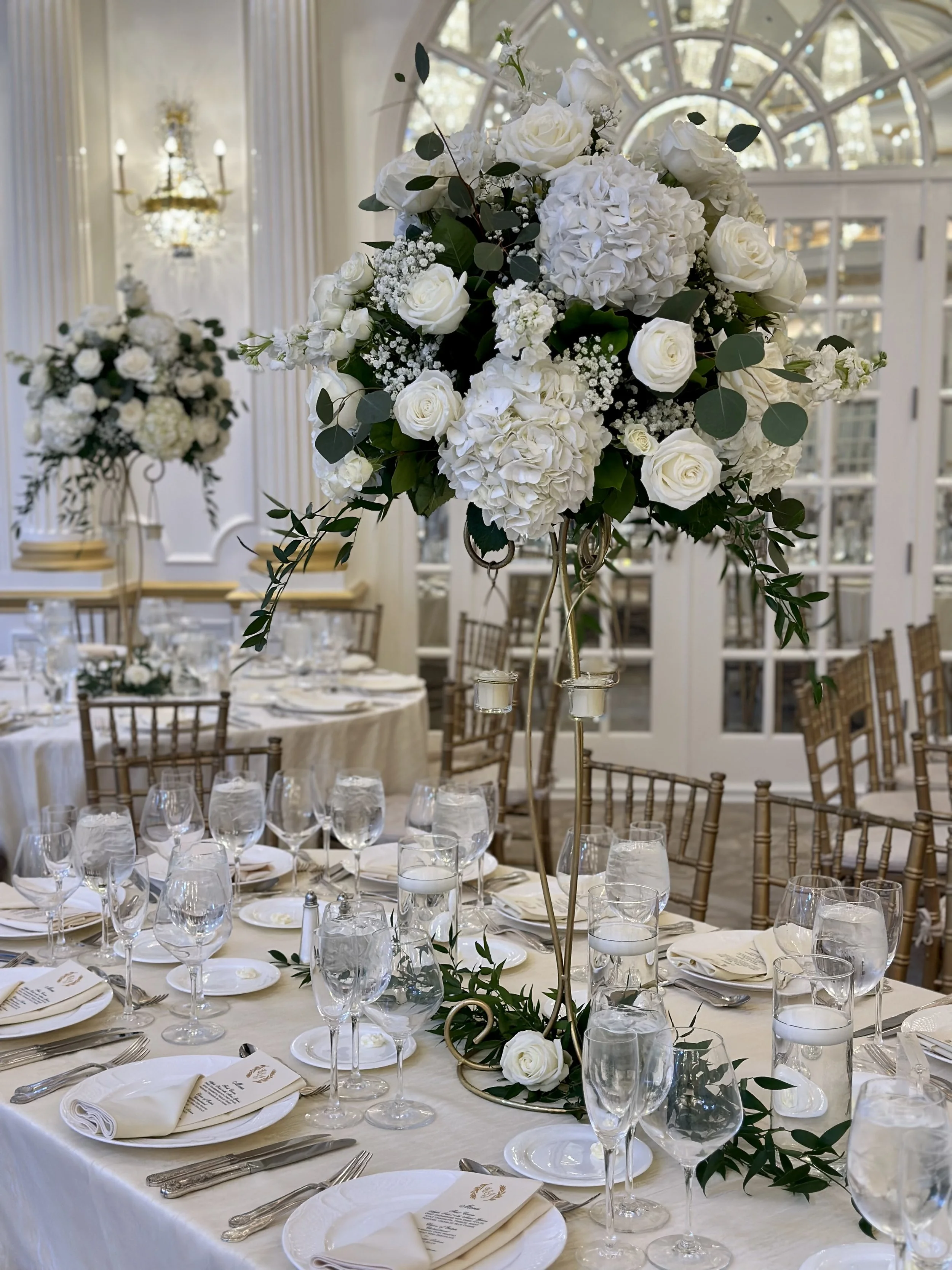 Elegant wedding reception table with a tall floral centerpiece of white roses, hydrangeas, and greenery, set in a bright room with large windows and a chandelier.