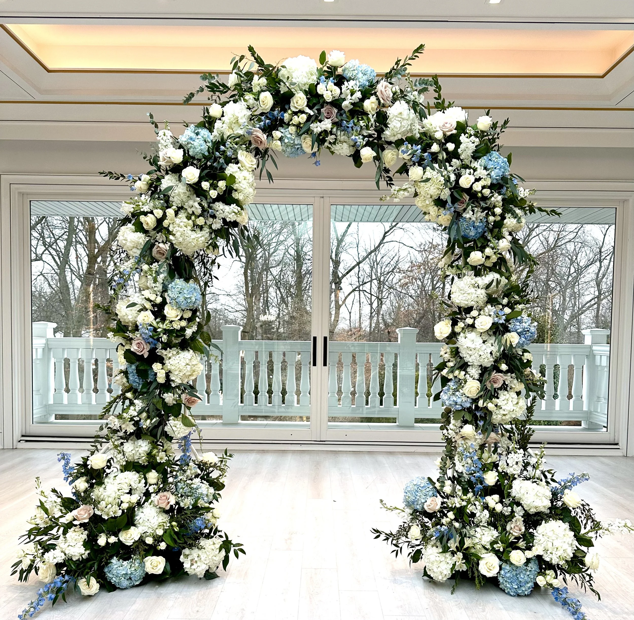 A large floral arrangement arch made of white, blue, and blush pink flowers and green foliage, set indoors in front of a glass door with a wooded outdoor view.