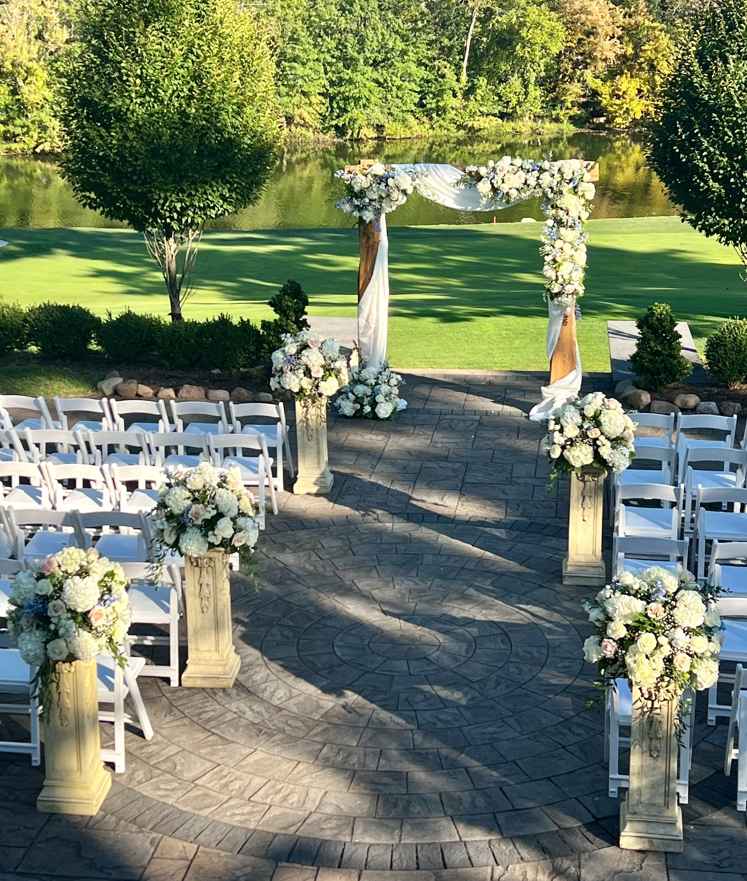 A beautiful outdoor wedding ceremony setup with an arch decorated with white and pastel flowers, positioned by a pond with lush green trees in the background.
