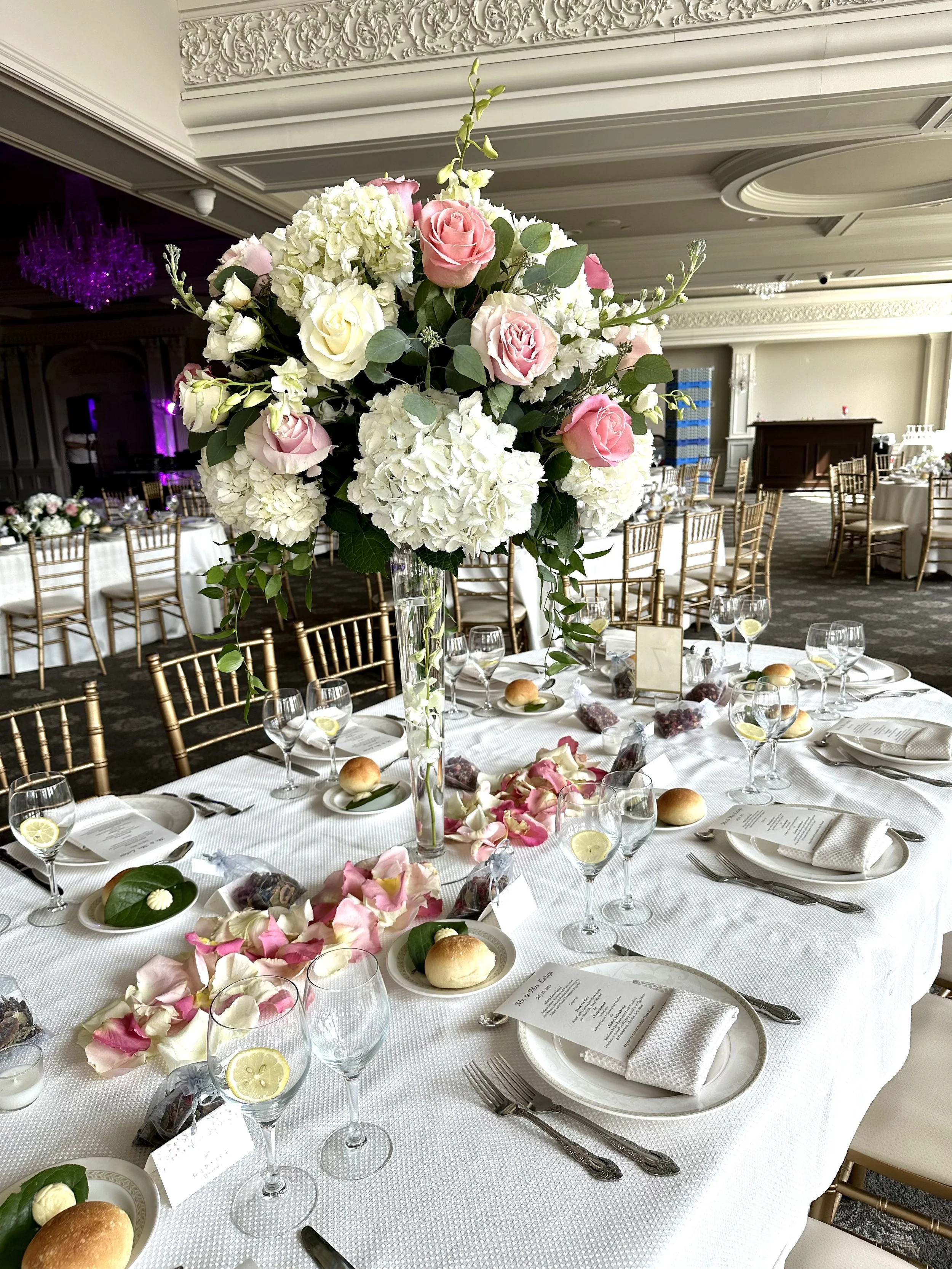 Wedding reception table with a large floral centerpiece of white hydrangeas and pink roses, set with plates, glasses, silverware, a menu, bread rolls, and scattered flower petals in an elegant decorated banquet hall.