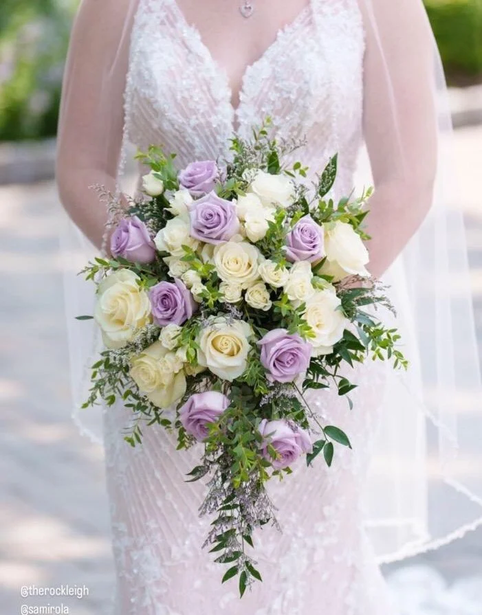 A bride holding a cascading bouquet of white and lavender roses with greenery, wearing a white lace wedding dress with sheer sleeves.