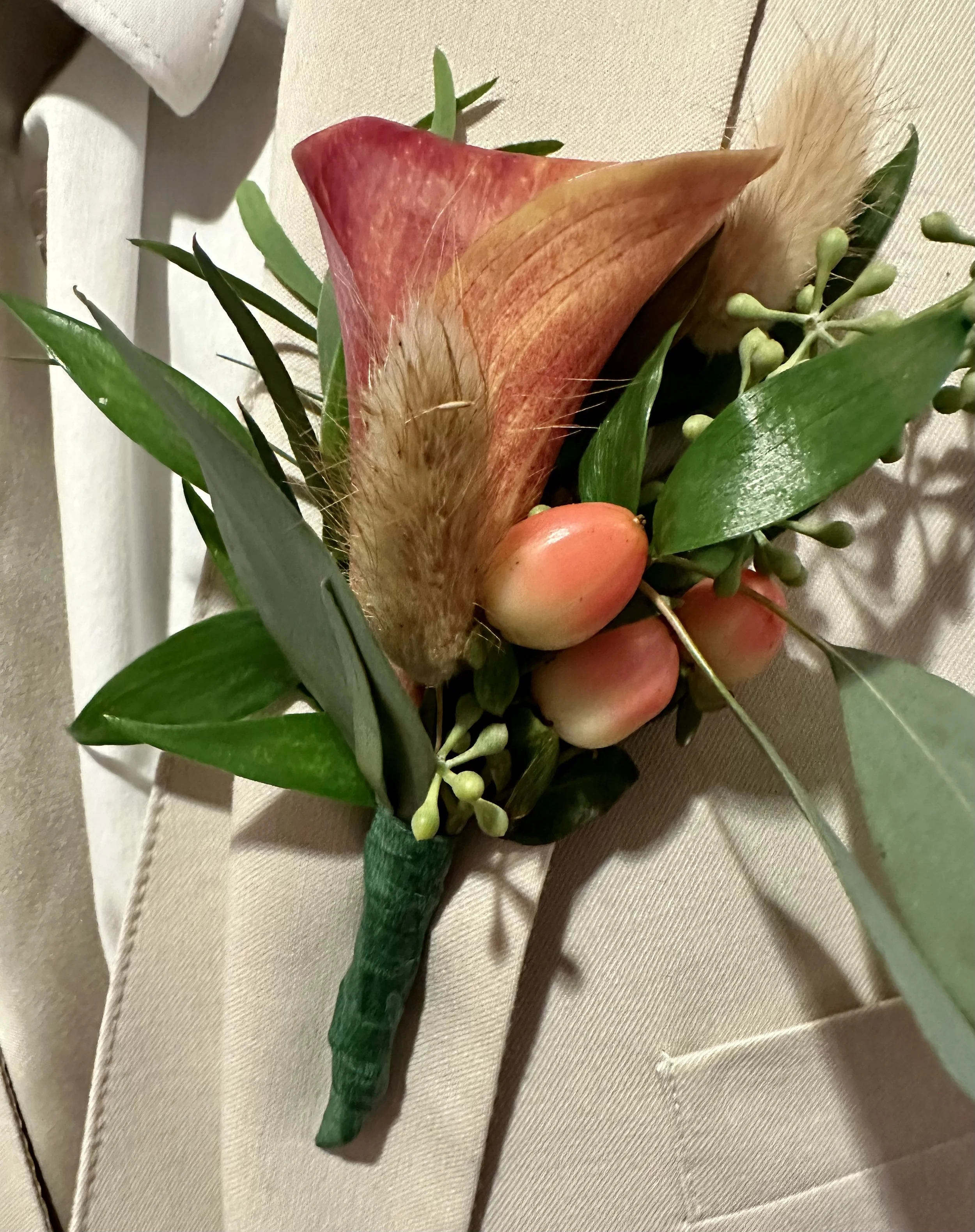 A boutonniere featuring a pink rose, greenery, and a beige feather, tied with a green ribbon, resting on a beige fabric surface.