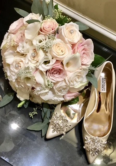A bouquet of pink and white roses with white calla lilies and greenery, paired with elegant cream-colored designer high heels adorned with rhinestone embellishments.