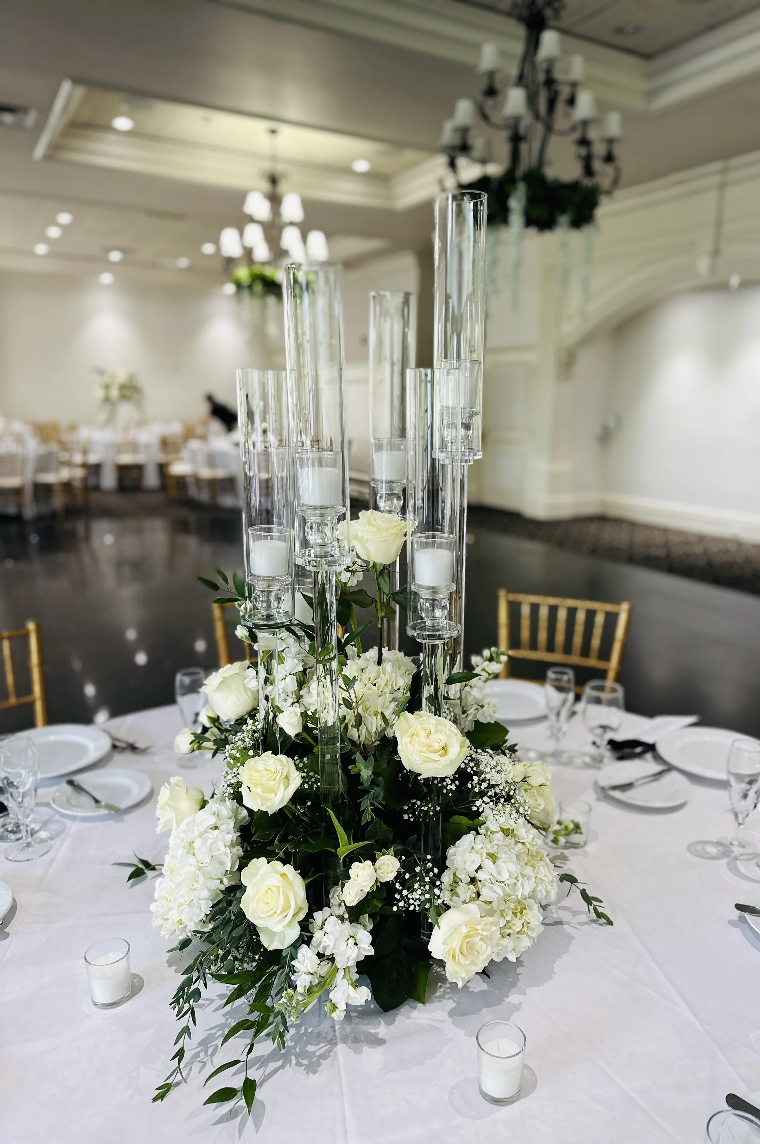 Elegant floral centerpiece with white roses and cascading greenery on a round table decorated for a formal event, with white tablecloths, gold chairs, and a banquet hall setting in the background.
