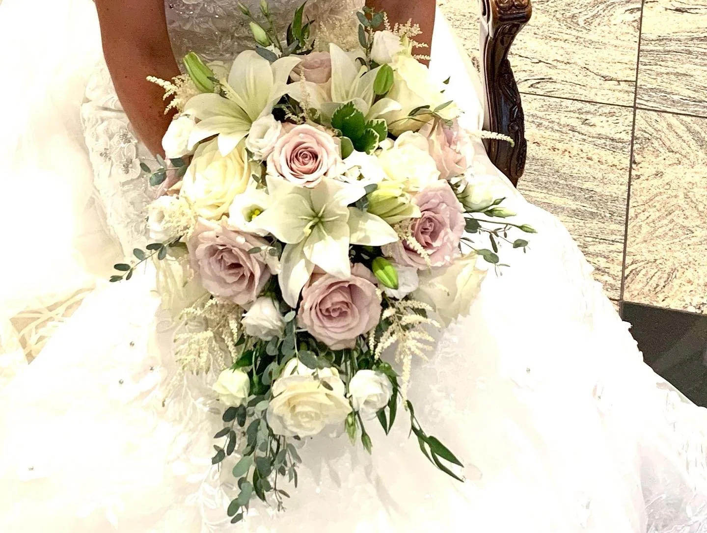 A bridal bouquet with white lilies, pale pink roses, white roses, and greenery resting on a white dress in front of a wooden chair.