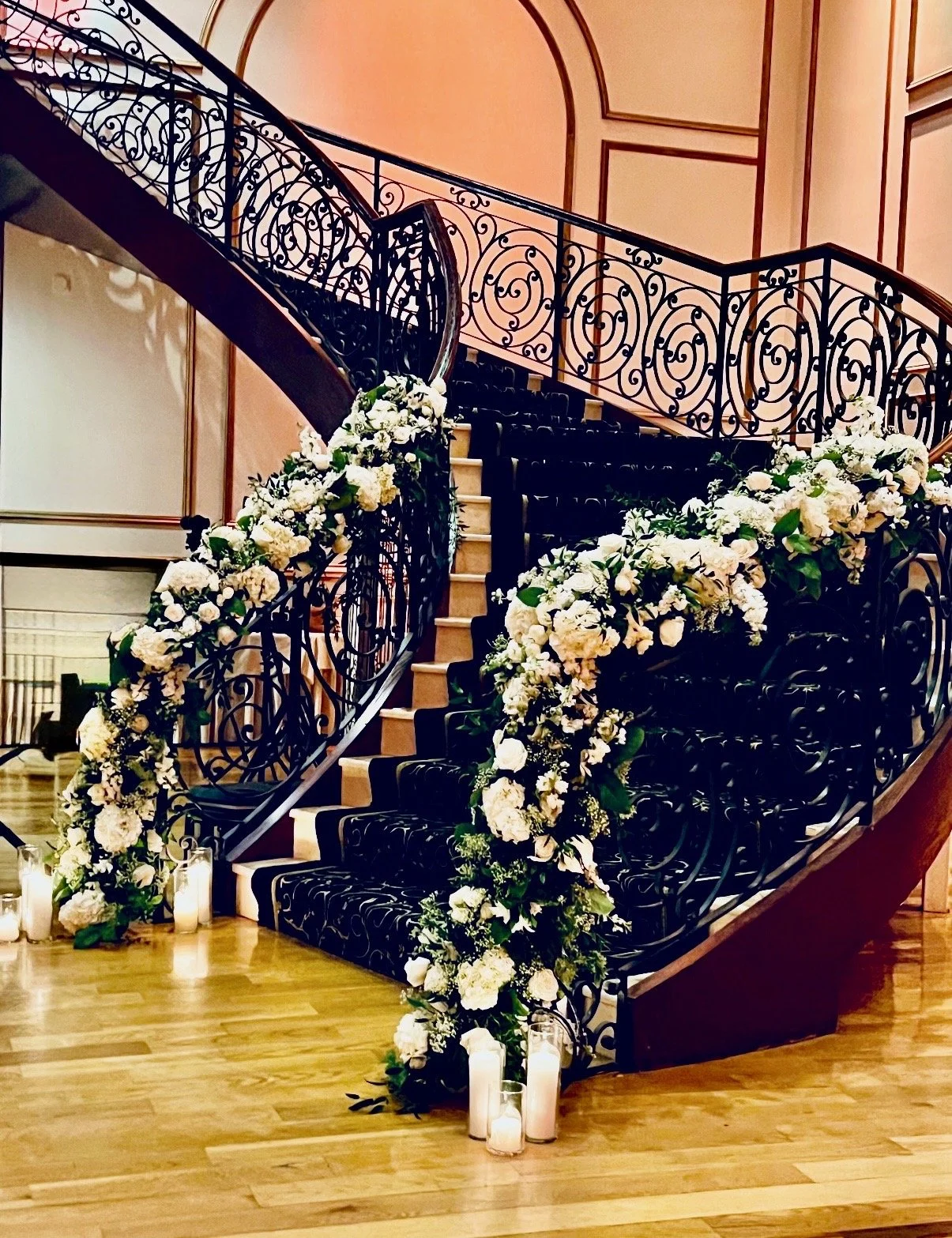 Elegant staircase decorated with white flowers and candles, likely for a wedding or special event, with pink walls and wood flooring.