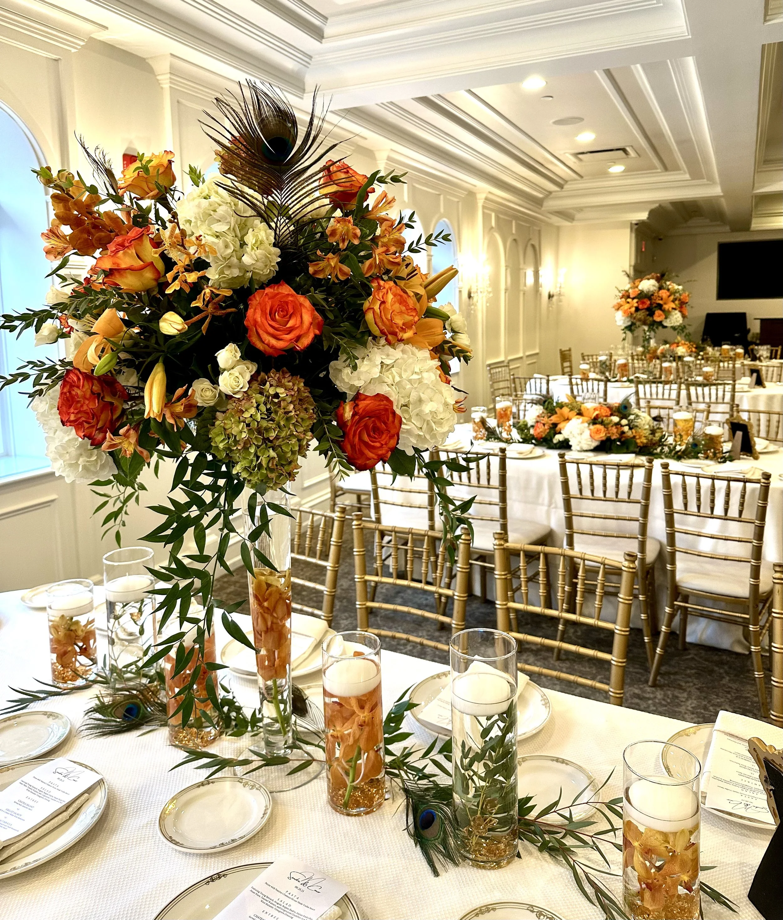 Elegant banquet table setup with a floral centerpiece featuring orange, white, and yellow flowers, accented with peacock feathers. The table is decorated with candles in glass holders, and place settings with menus. In the background, additional tabl