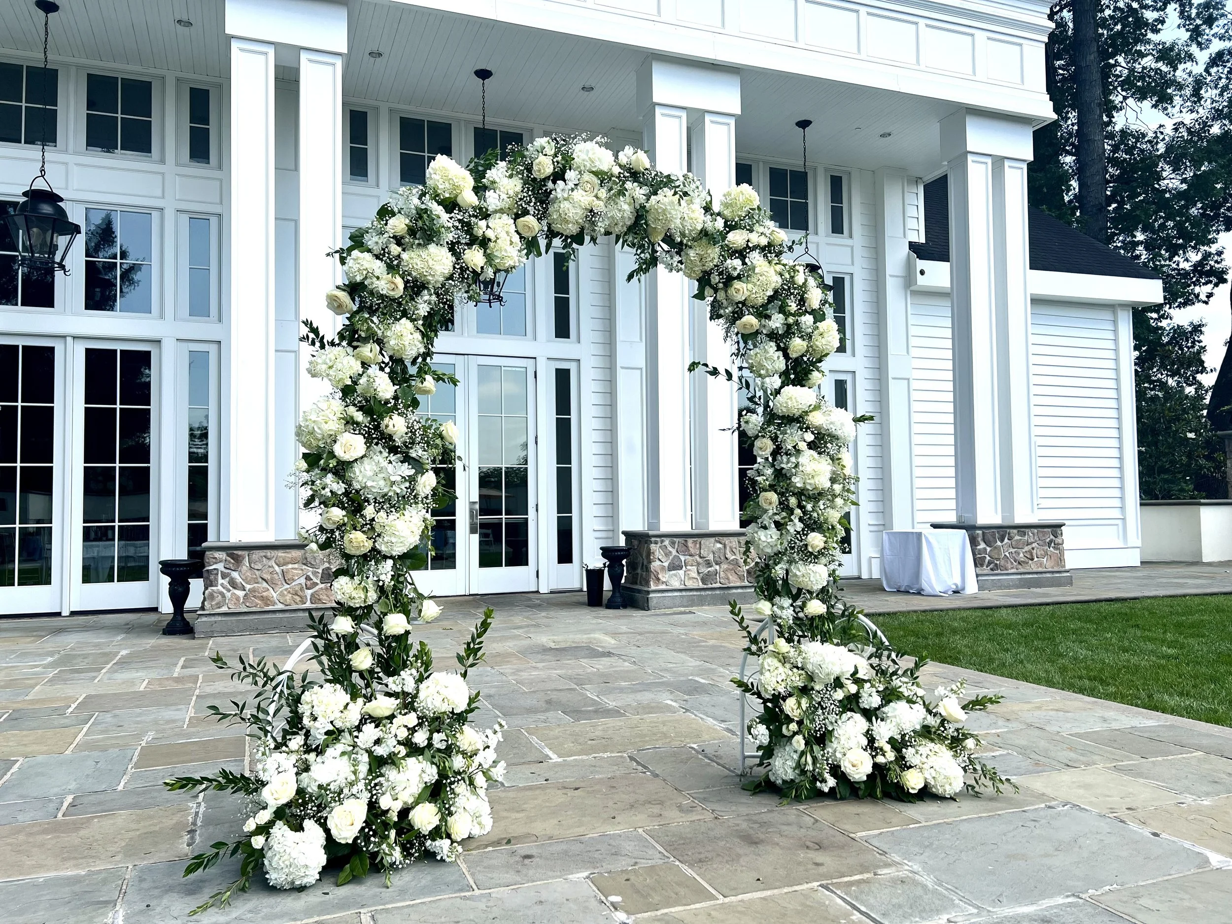 White floral arch and floral arrangements on the ground outside a white building with large windows and a stone foundation, likely set up for a wedding ceremony.