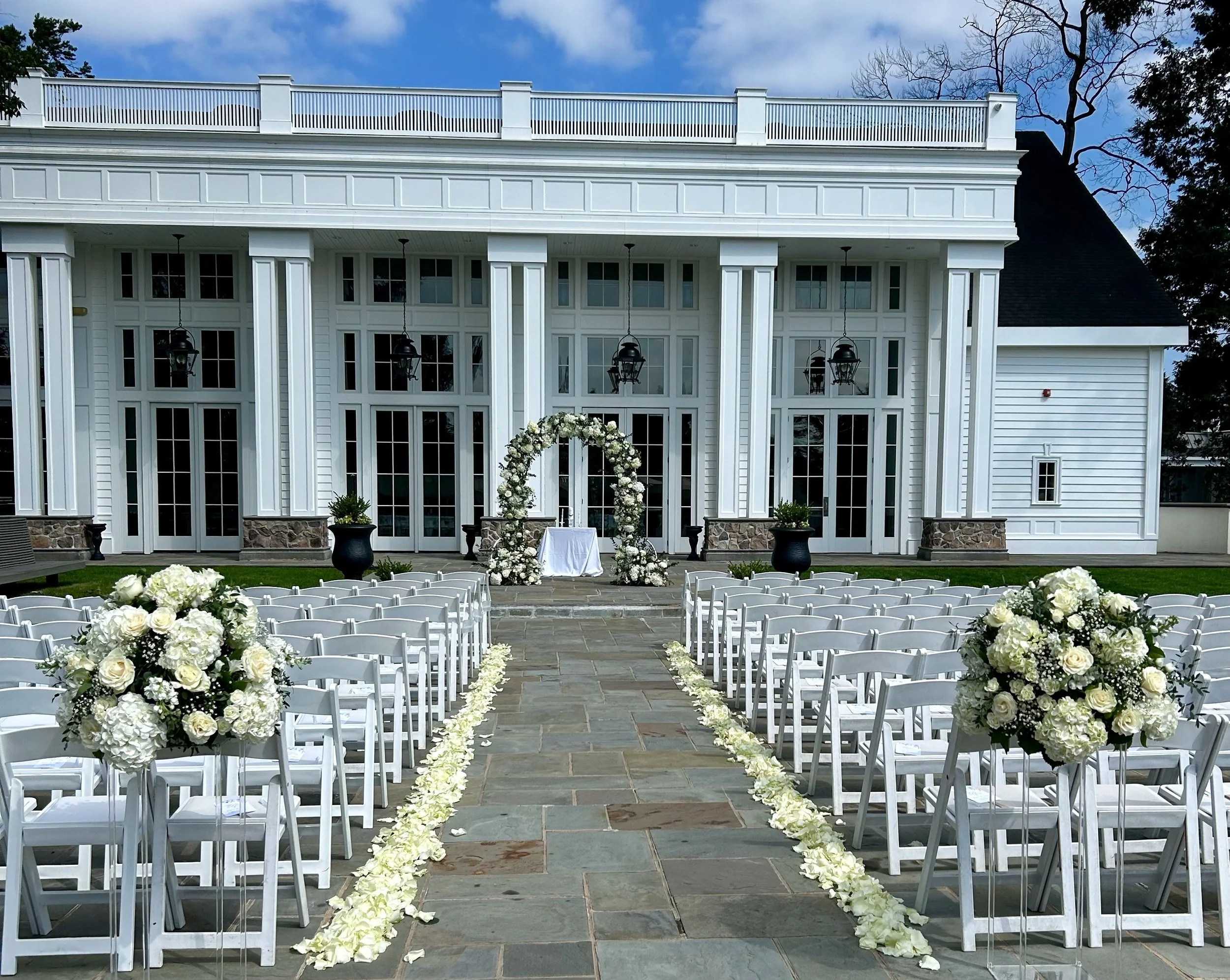 Outdoor wedding setup with white chairs arranged in rows on either side of a flower-decorated aisle, leading to a floral arch against a large white house with columns and a balcony, under a partly cloudy sky.