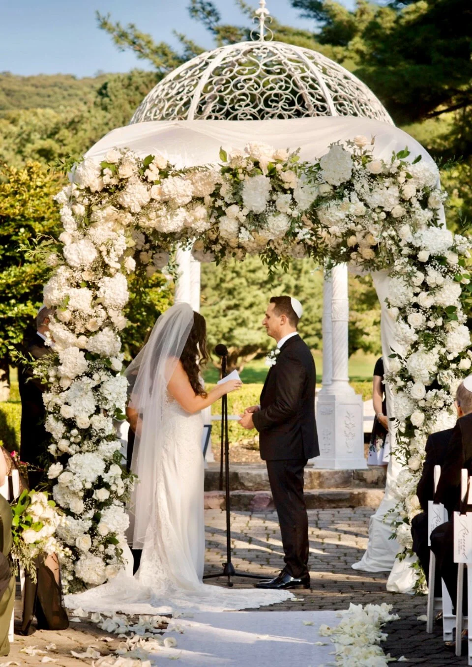 A wedding ceremony outdoors with a bride and groom standing under a floral arch decorated with white flowers and greenery, exchanging vows. The bride wears a white gown and veil, while the groom is in a black suit with a white boutonniere. The area i