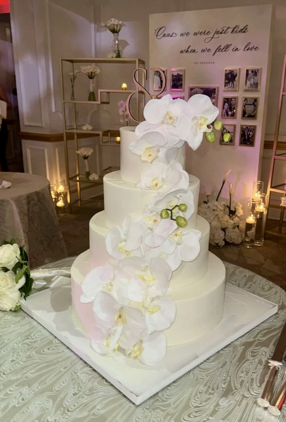 A multi-tiered wedding cake decorated with cascading white orchids and green buds, topped with a pink 'S' monogram, positioned on a silver tray at a festive event.