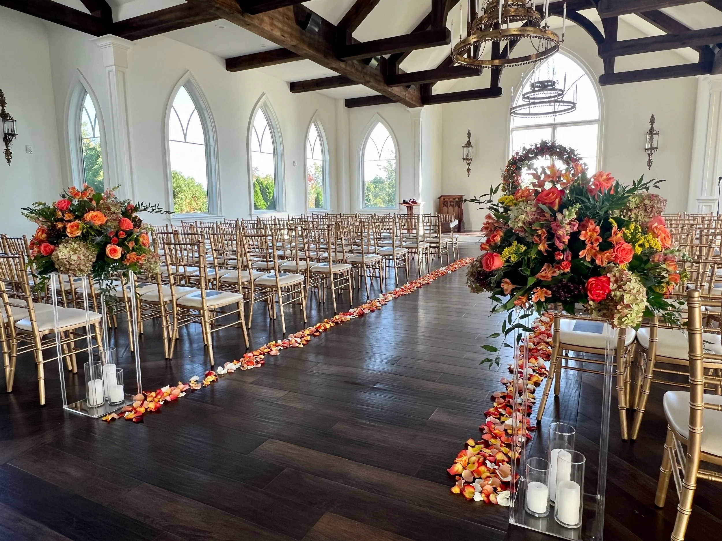 Interior of a wedding venue with rows of golden chairs, large flower arrangements with pink, peach, and white flowers at the entrance, and rose petal aisle decorations on a wooden floor. Large arched windows let in natural light.