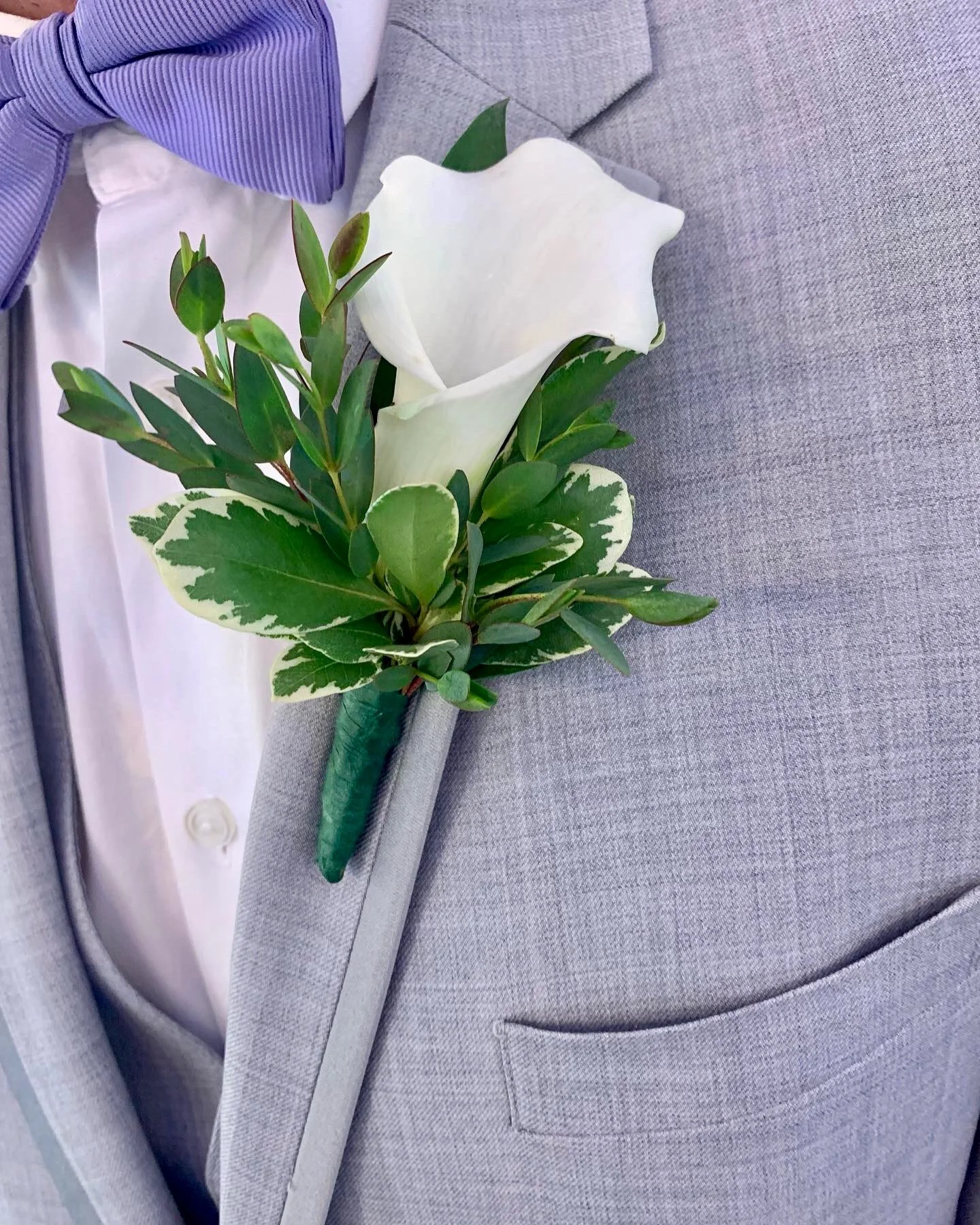 A close-up of a gray suit jacket with a white shirt and a purple striped bow tie. On the lapel, there is a boutonniere with a white calla lily and green leaves.