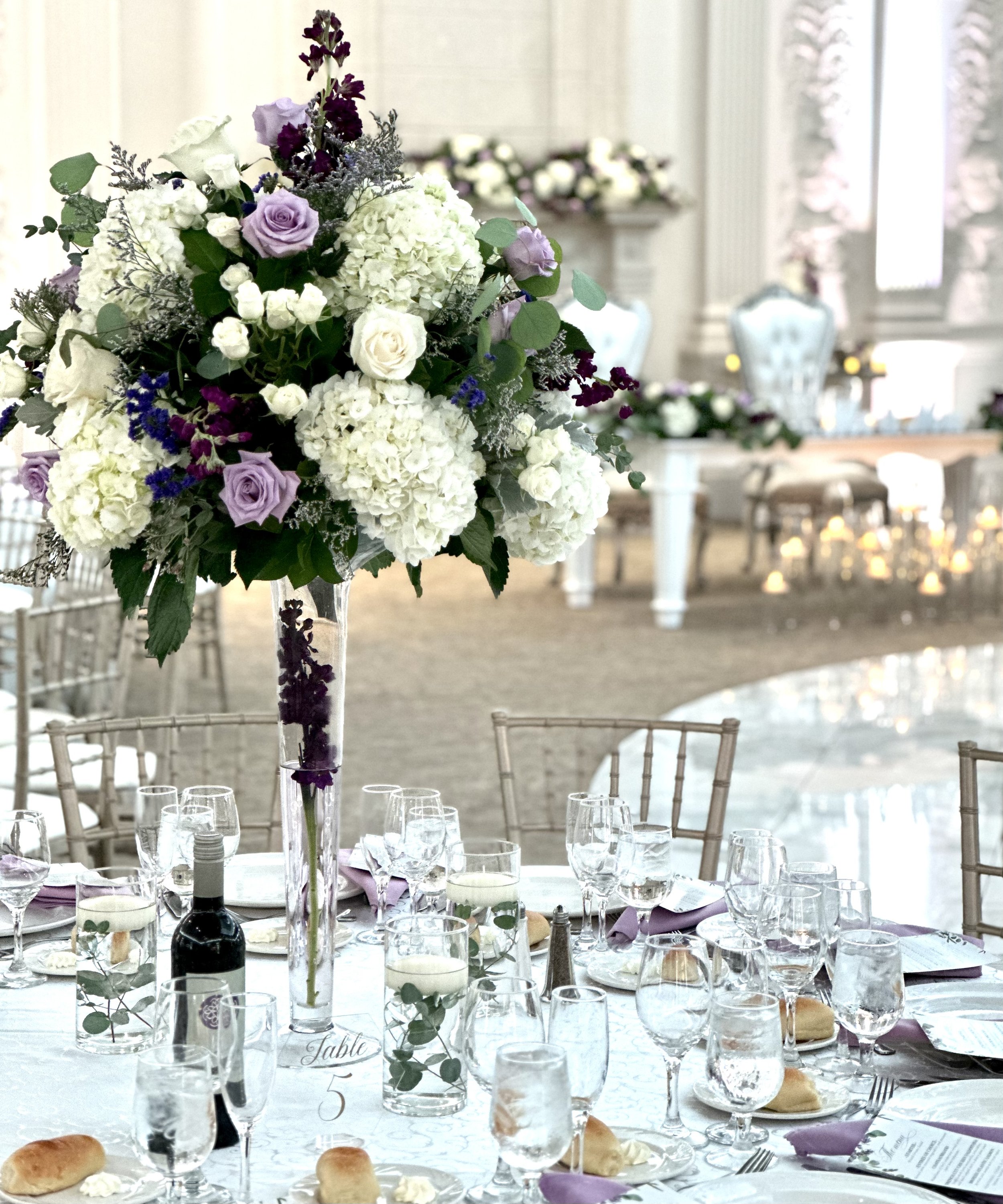 A formal event setup with a large floral centerpiece on a dining table. The centerpiece features white and purple flowers, including roses and hydrangeas, with greenery. The table is set with wine glasses, water glasses, bread rolls, and utensils. Th