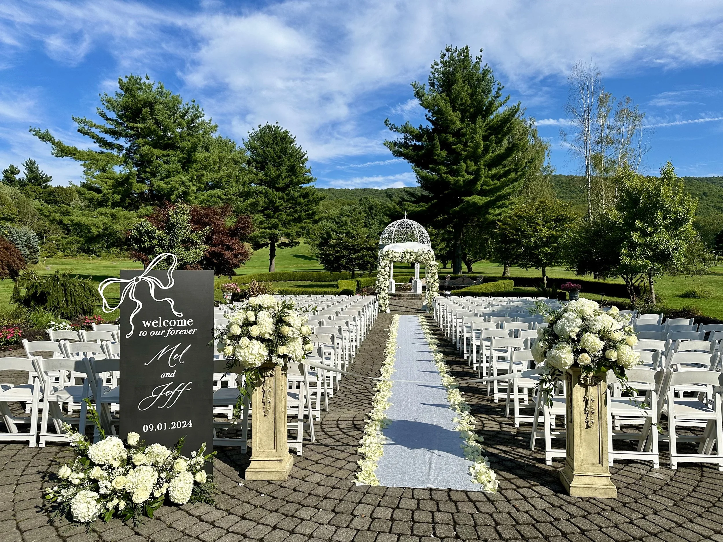 Outdoor wedding ceremony setup with white chairs, floral arrangements, and a wedding arch on a paved area surrounded by trees and mountains under a blue sky.