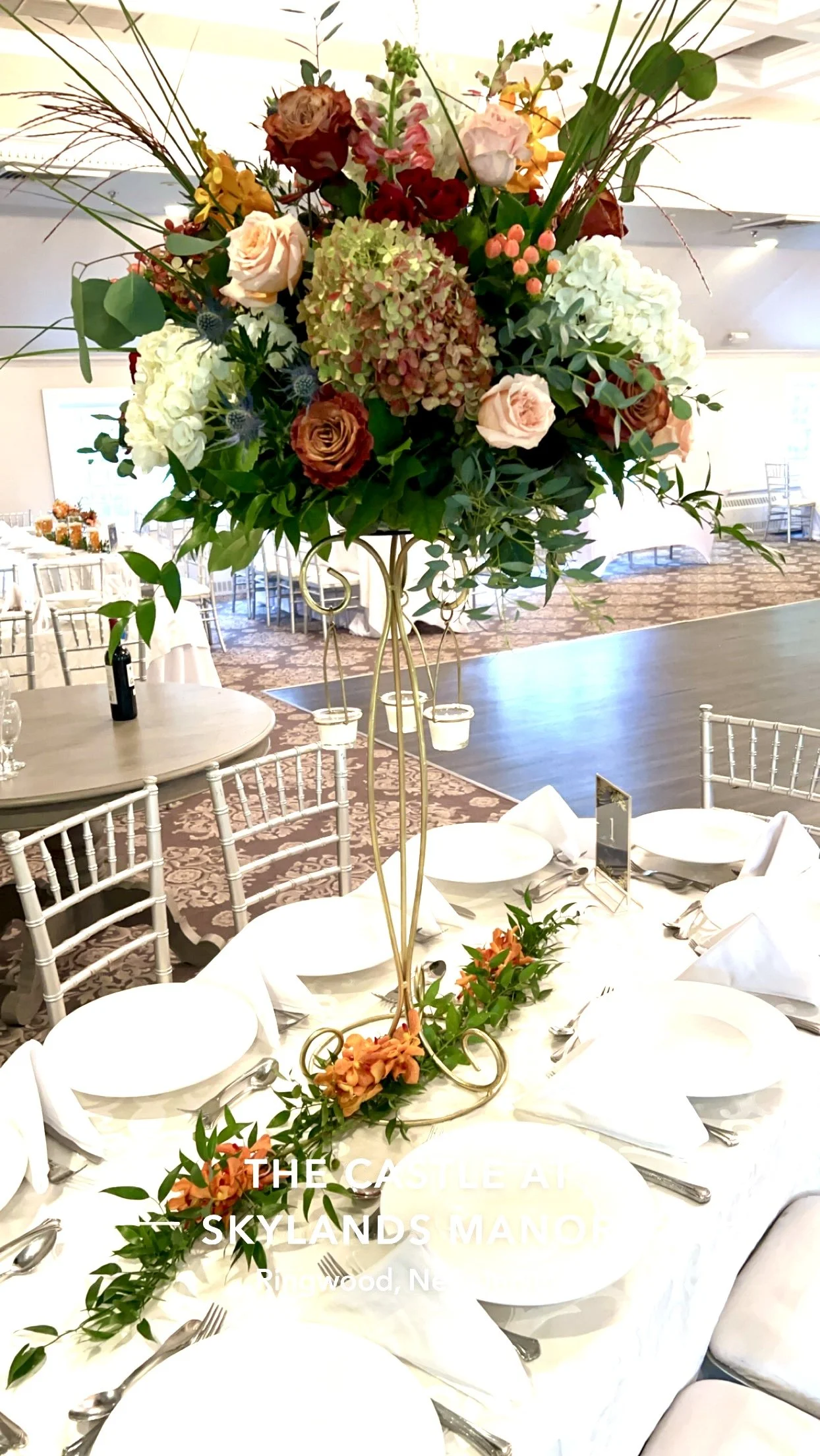 A tall floral centerpiece with pink and white roses, hydrangeas, and greenery on a banquet table at an event space.