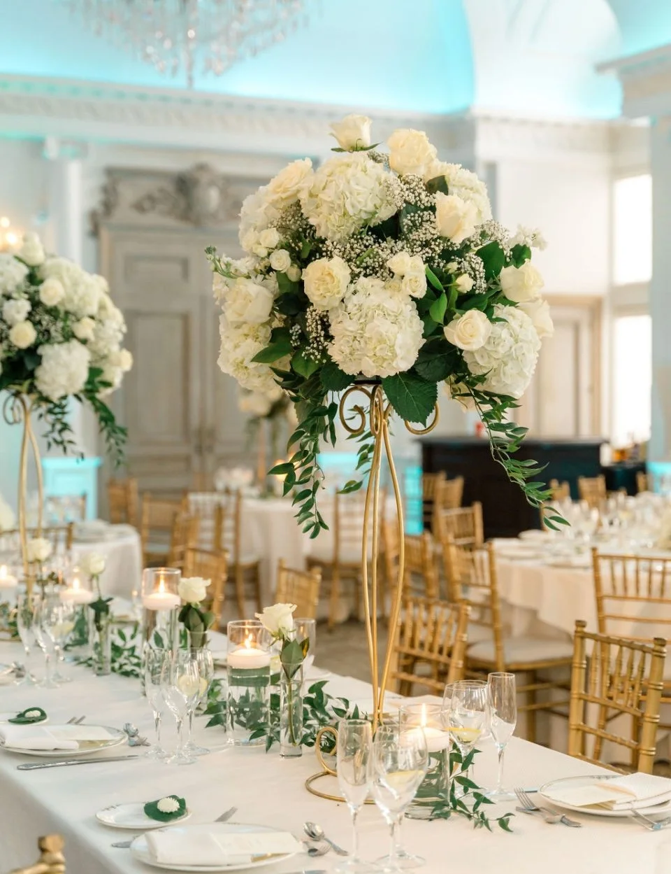 Wedding reception table decorated with white floral centerpieces, candles, and elegant table settings in a decorated ballroom.