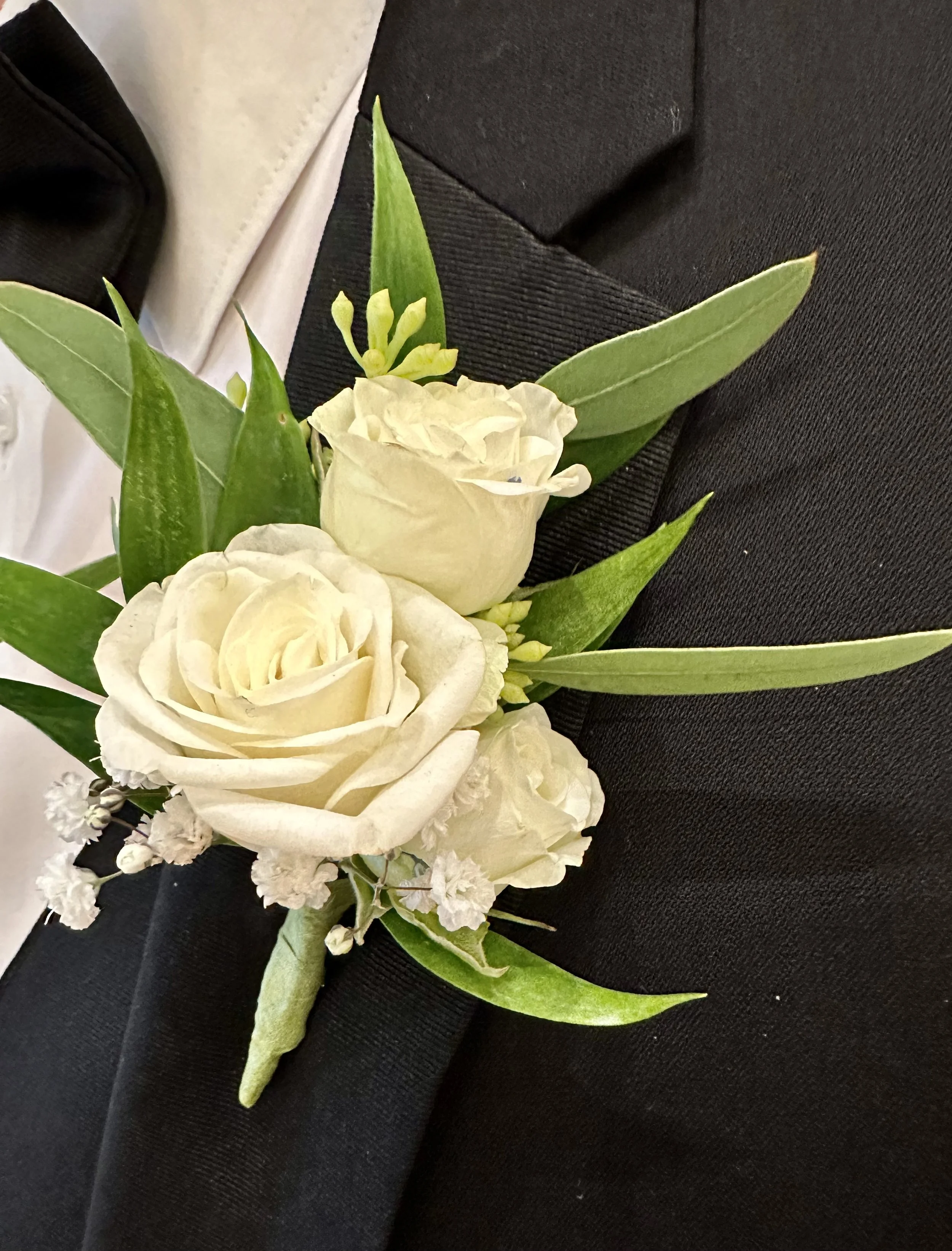 A boutonniere with white roses, greenery, and small white flowers, pinned to a black fabric background.