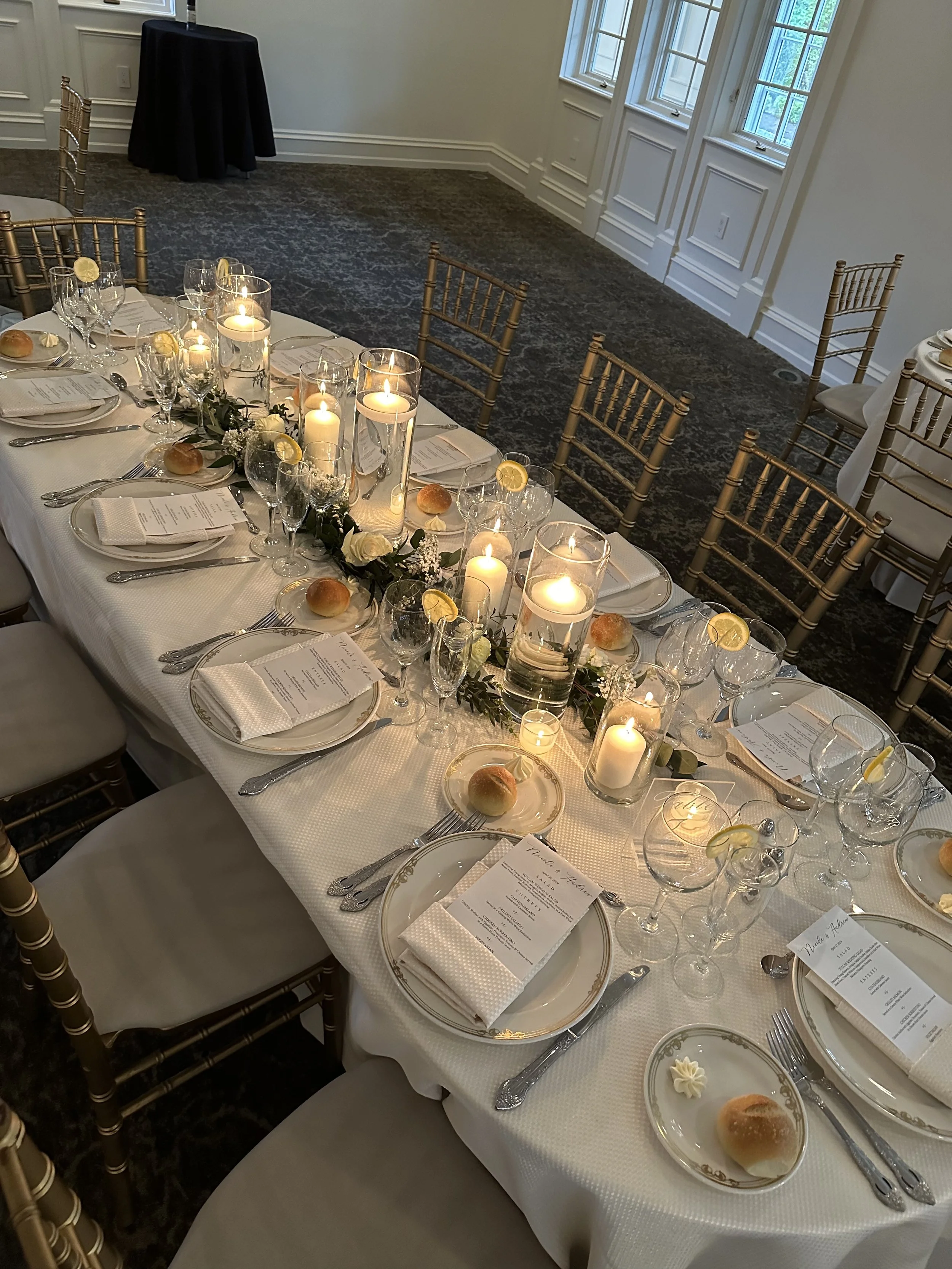 A decorated dining table set for a formal event, featuring candles, floral arrangements, bread rolls, menus, and neatly arranged glassware and silverware, with chairs around the table.