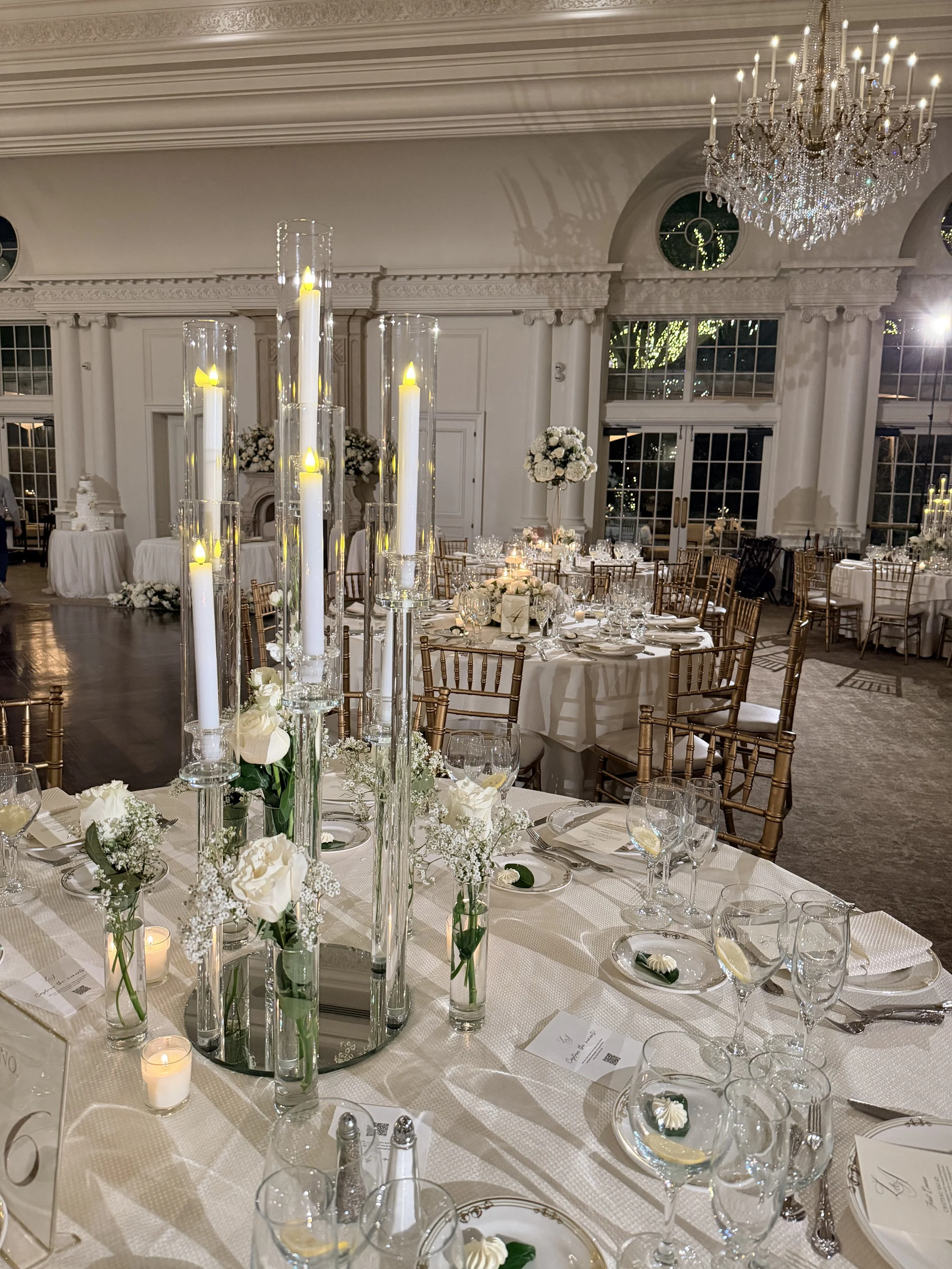 Elegant banquet hall decorated for a wedding reception with white floral arrangements, tall candles, and chandelier lighting.