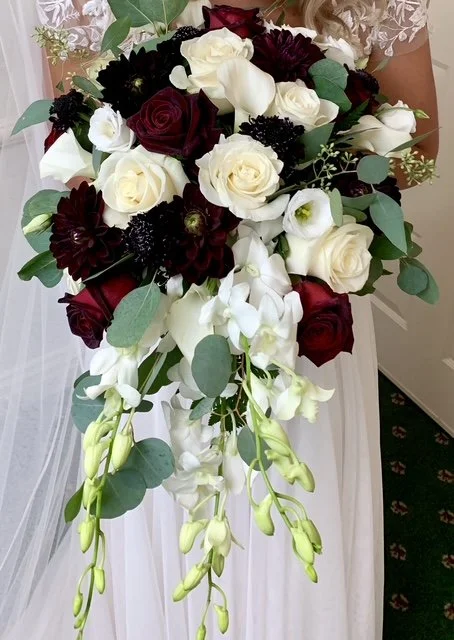 Bridal bouquet with white, red, and dark purple roses, surrounded by white orchids and greenery.