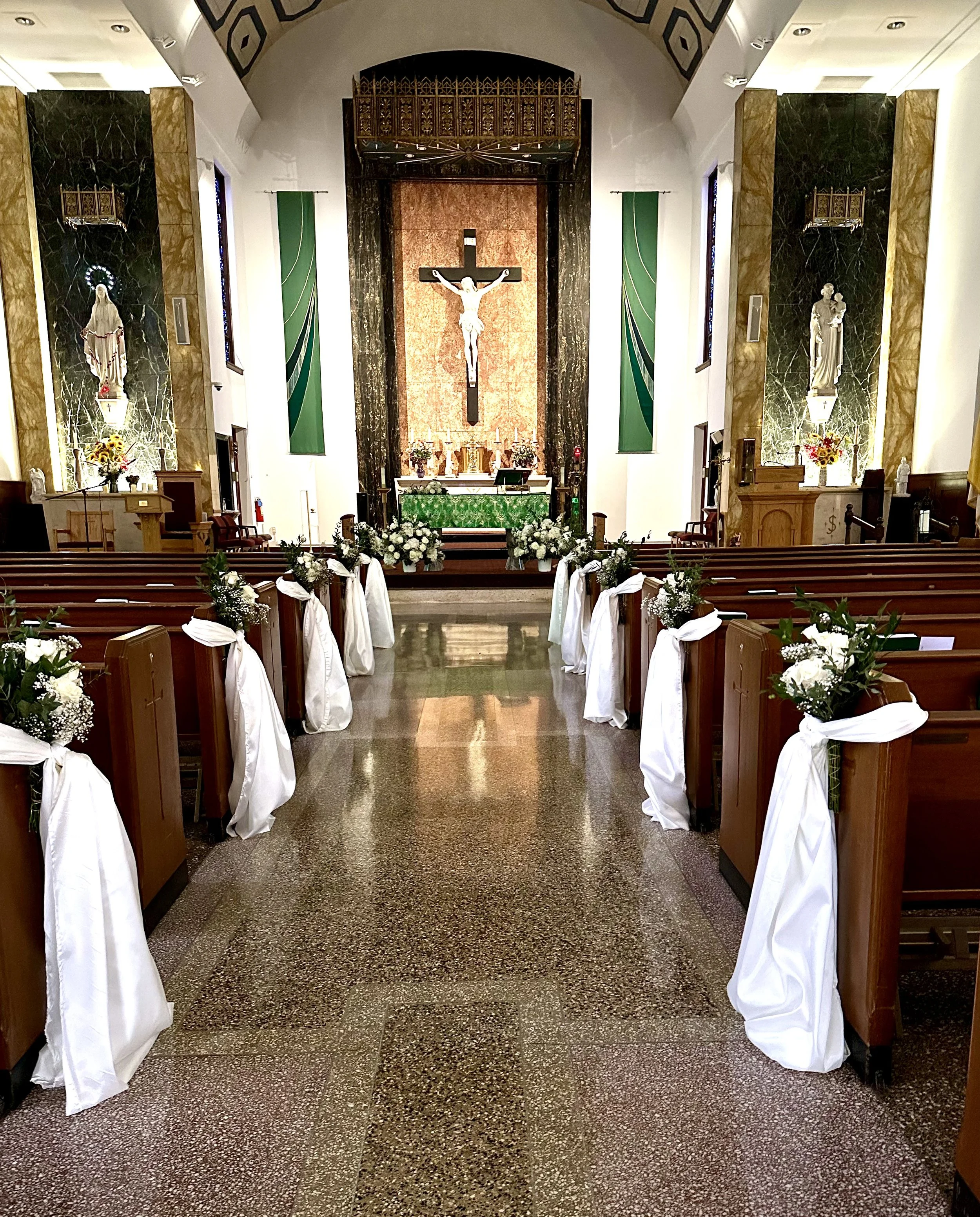 Interior of a church with an altar and pews decorated with white flowers and ribbons, featuring a crucifix on the wall behind the altar.