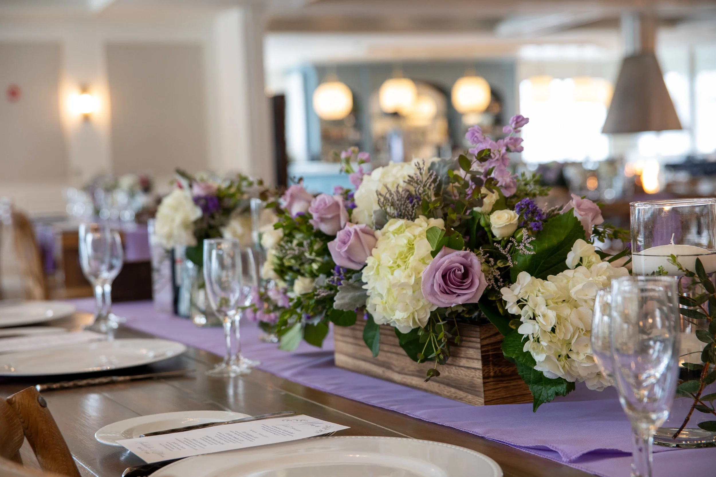 A table decorated with a floral centerpiece consisting of white, purple, and pink flowers in a wooden container, surrounded by wine glasses and white plates in an elegant indoor setting.