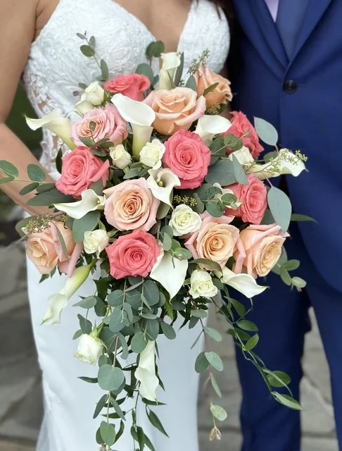 A bride in a white lace dress holding a cascading bouquet of pink, peach, and white roses, with white calla lilies and green foliage, standing next to a groom in a blue suit.